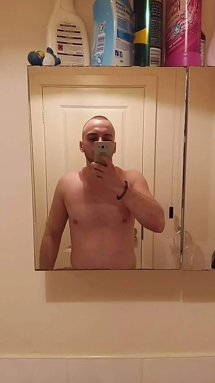 A shirtless man taking a selfie in a mirror, with a bathroom cabinet and cleaning products visible above him.