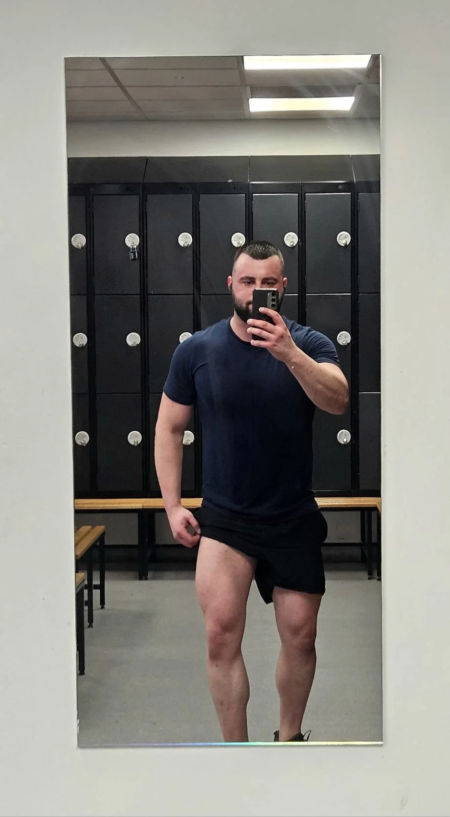 A man taking a selfie in a locker room, wearing a dark blue T-shirt and black shorts, with a beard and buzz cut hairstyle. He is standing in front of a mirror with black lockers behind him.