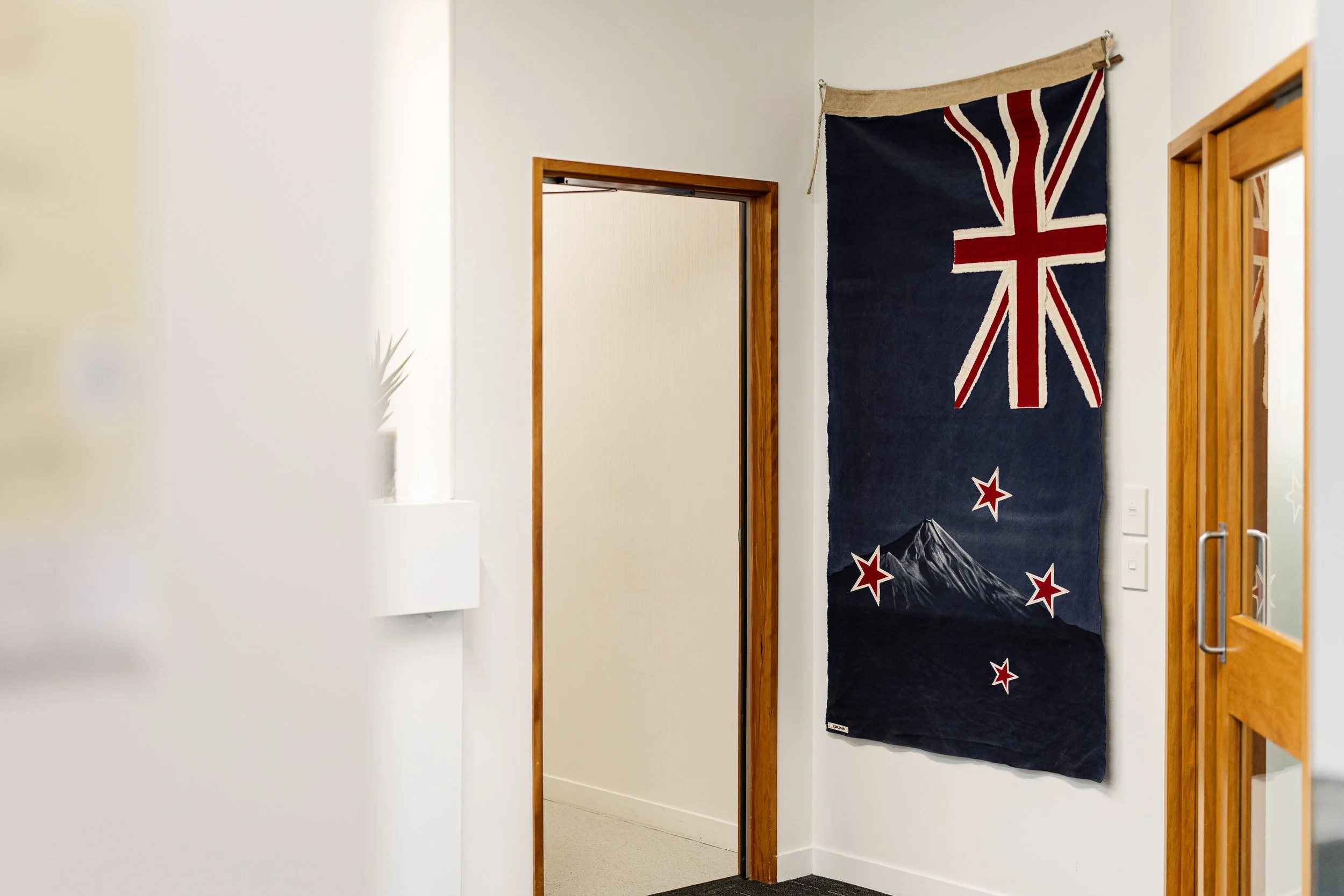 Interior of a room with a wooden doorway, white walls, and a decorative textile hanging with a Union Jack design and New Zealand mountains, stars, and flags.