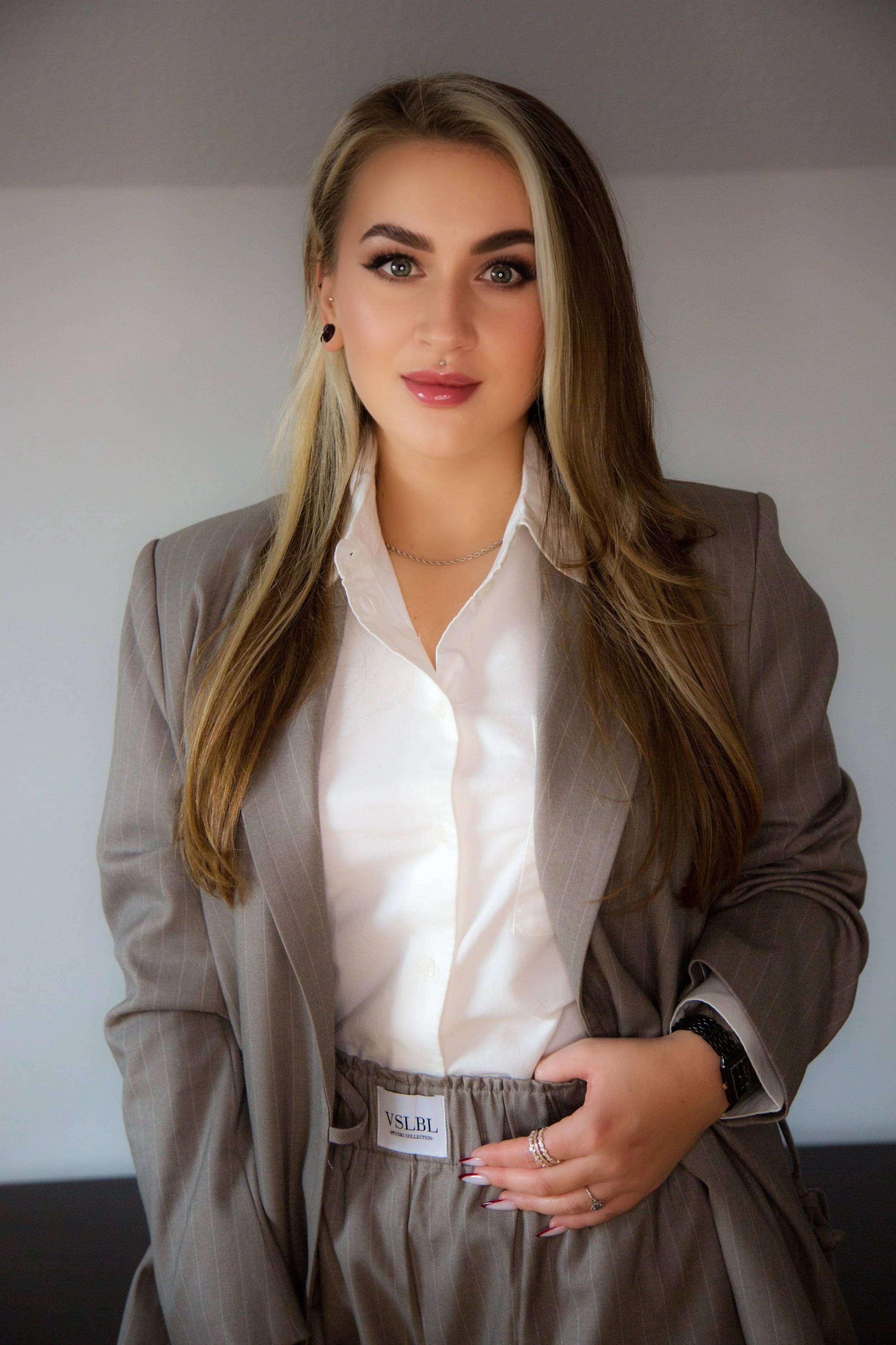 Junge Frau mit langen braunen Haaren, im grauen Anzug und weißem Hemd, posiert vor neutralem Hintergrund.