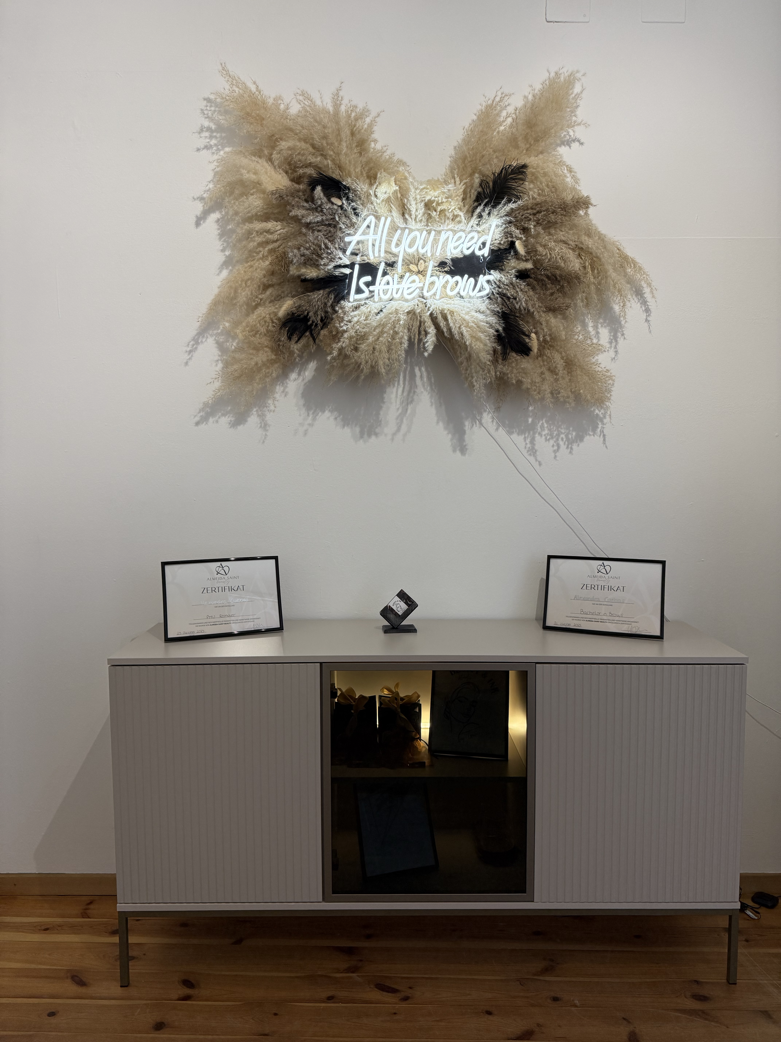 Kunstinstallation mit einem weißen Slogan-Leuchtschild, das die Worte 'All you need is love braws' zeigt, auf einer Wand über einem weißen Sideboard, mit Zertifikaten und kleinen dekorativen Gegenständen darauf, in einem modernen Raum mit Holzboden.