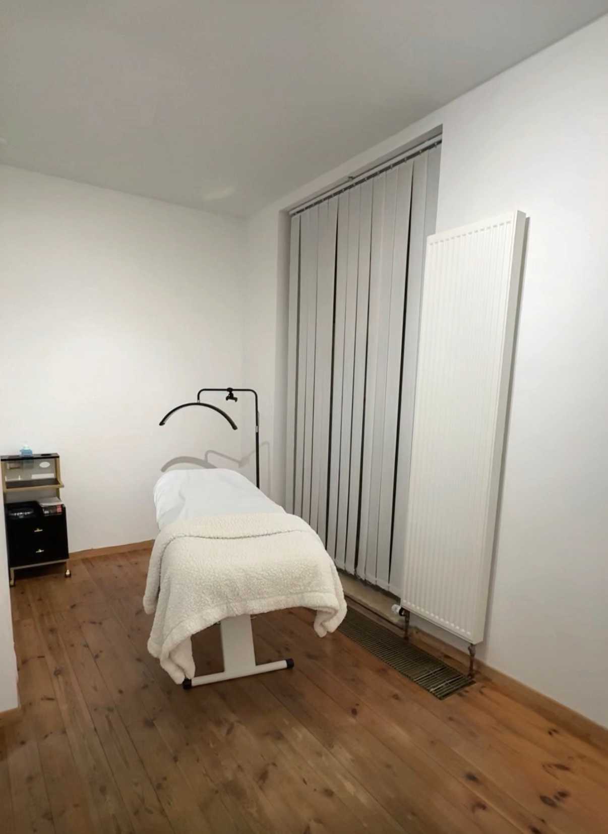 Behandlungsraum mit Behandlungsliege, kleinem Regal, medizinischer Geräte und Heizkörper an einer weißen Wand, Holzboden.
