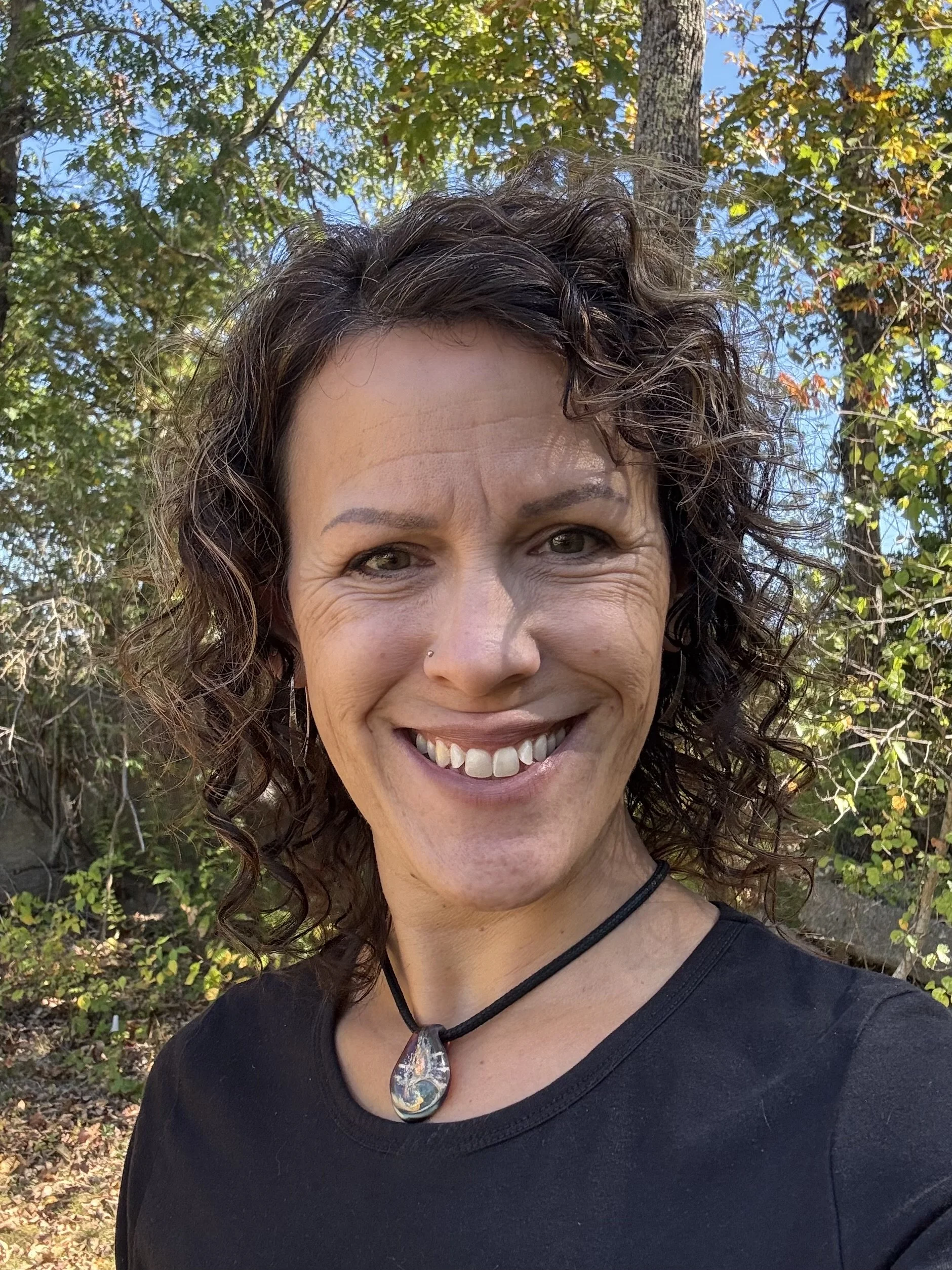 A woman with curly brown hair and a nose piercing smiling outdoors, wearing a black shirt and a teardrop-shaped pendant necklace, with trees and blue sky in the background.