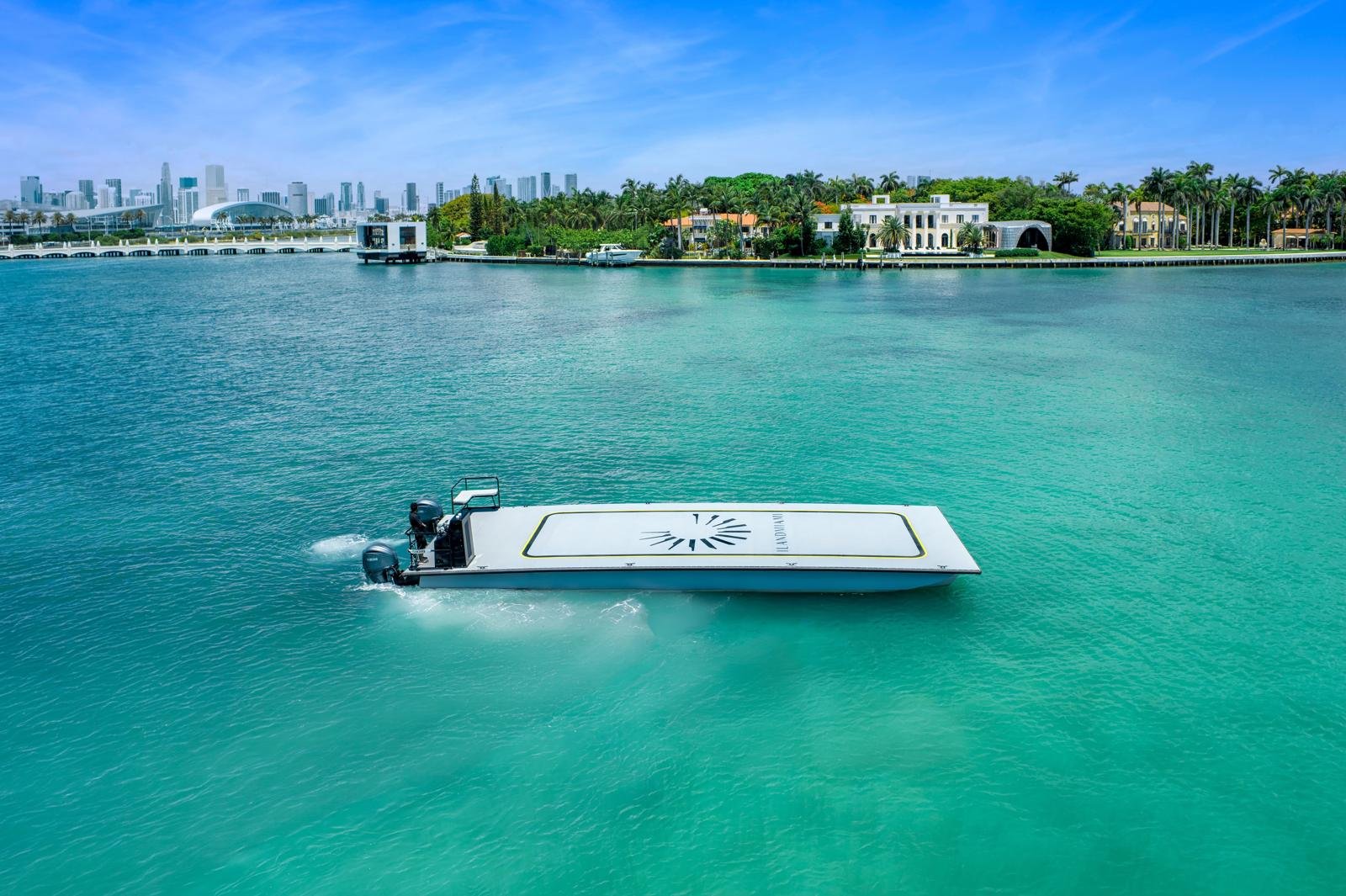A futuristic, sleek electric boat cruising on turquoise water with a city skyline and residential homes with palm trees in the background.