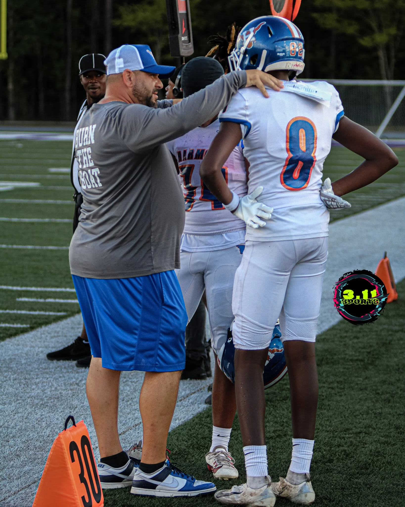 A football coach with a beard and blue cap giving instructions to a young football player wearing a blue helmet and white jersey, number 8, on a football field during a game. Two other players and a referee are in the background. An orange marker with the number 30 is on the sideline.