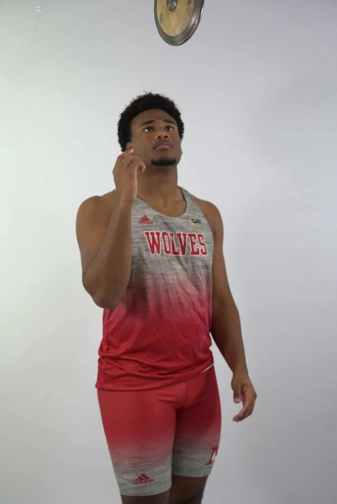 A male athlete wearing a red, gray, and white sports uniform with "WOLVES" written on the front, standing and looking up at a clock on the ceiling.