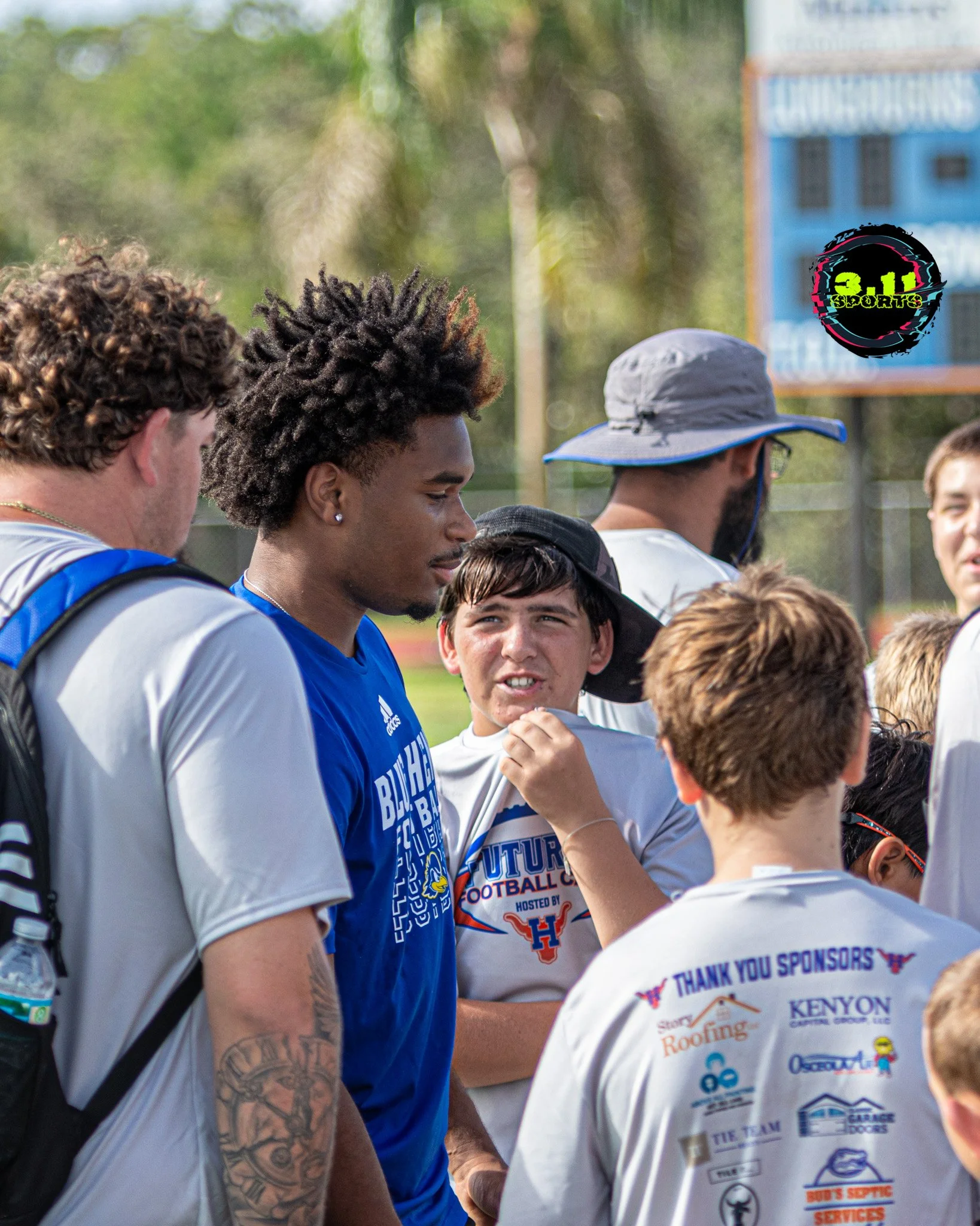 A group of football players gathered outdoors on a sports field, with some wearing team shirts and others in casual clothing, engaging in a discussion, with a scoreboard visible in the background.