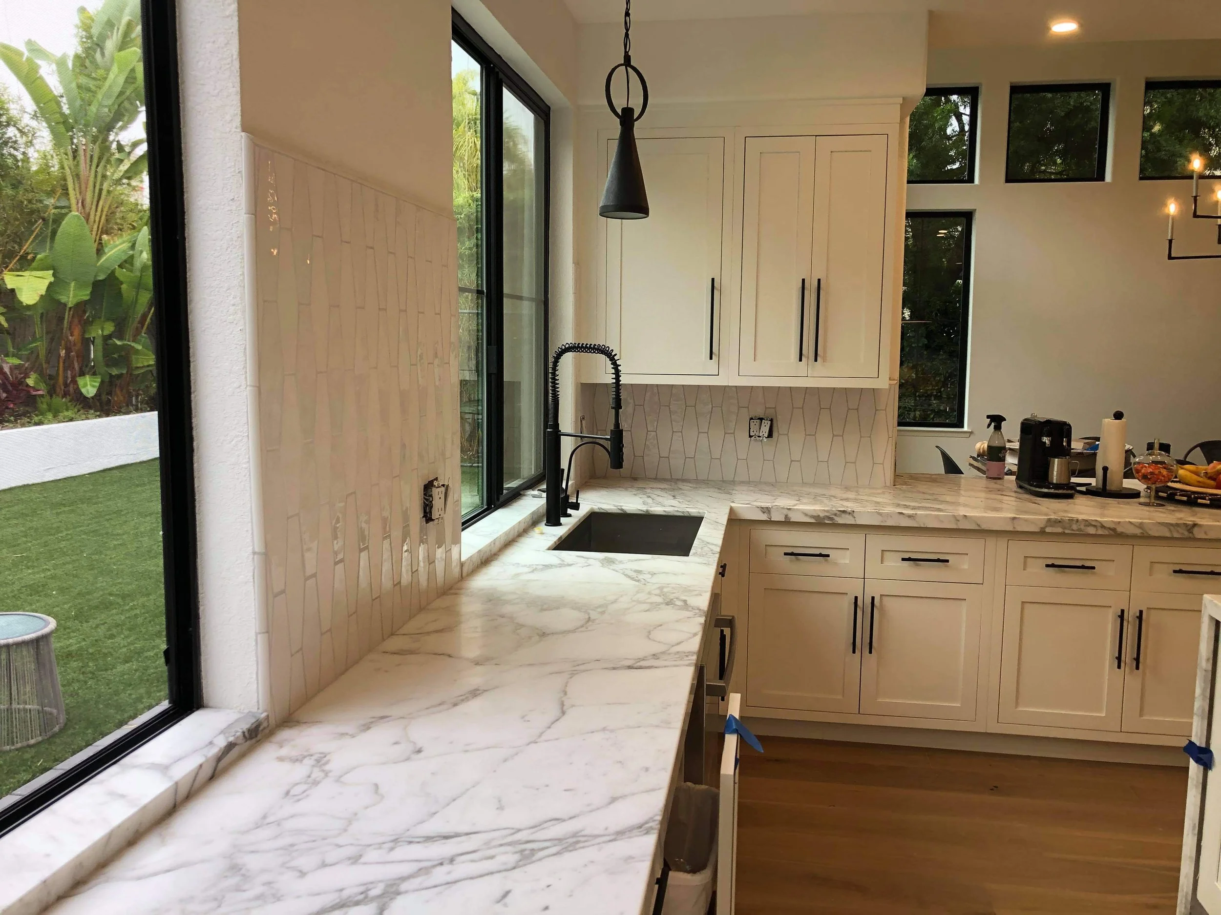 Modern kitchen with marble countertops, white cabinets, black fixtures, and large windows overlooking a garden.