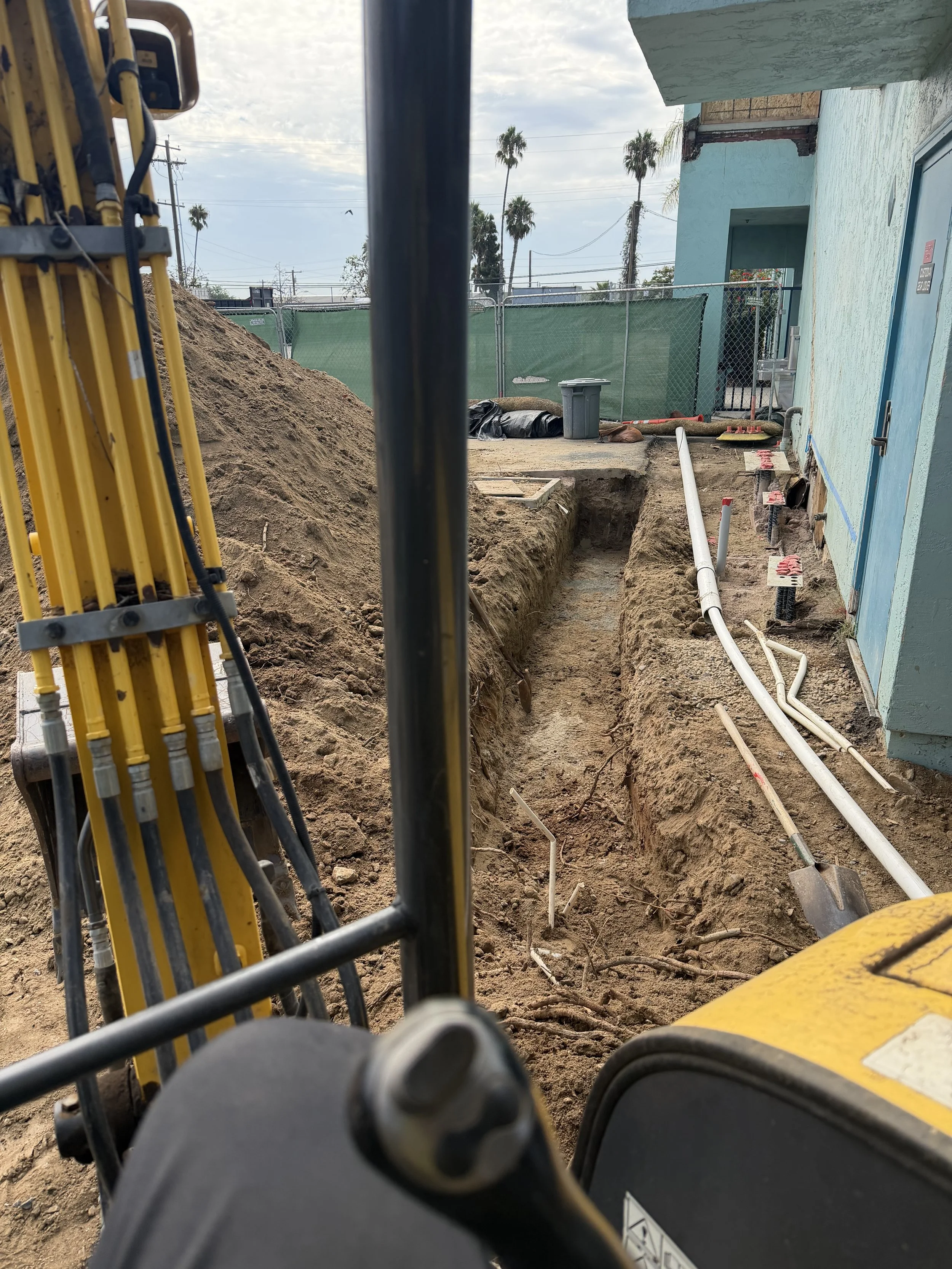 Construction site with a trench dug beside a light blue building, pipes installed along the trench, construction equipment, and palm trees in the background.