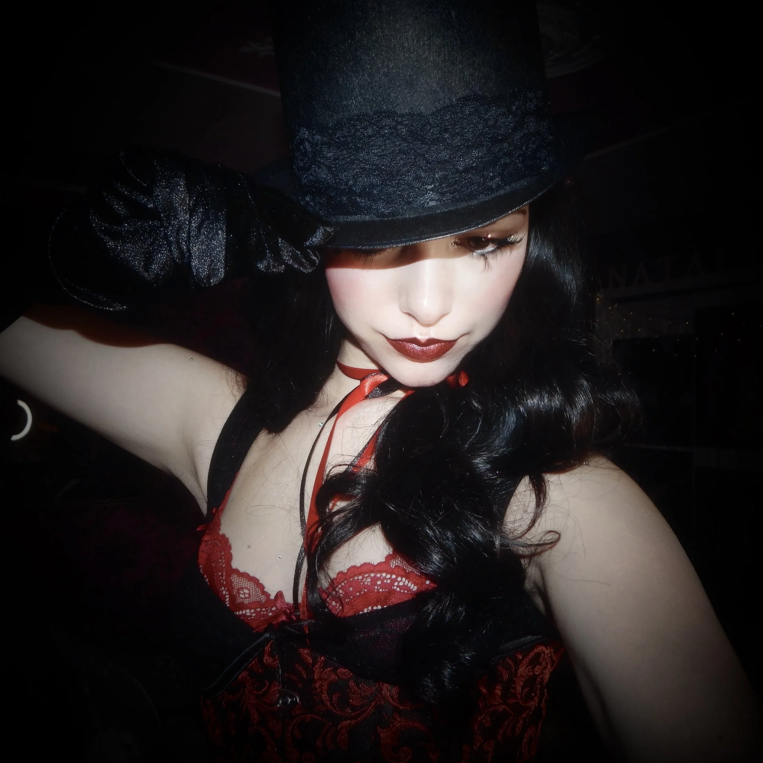 A woman with long, black, wavy hair wearing a black top hat, black outfit with red lace accents, and black gloves, posing with her head tilted downward.