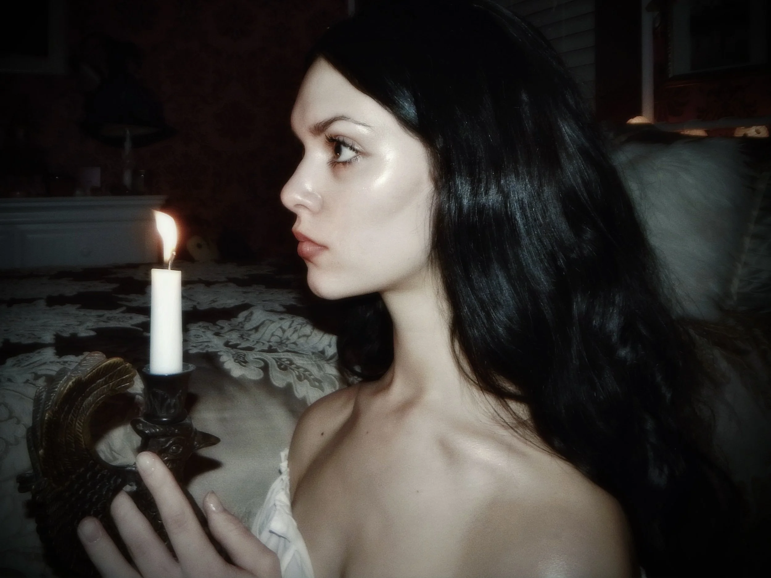 A woman with long black hair and fair skin holding a lit candle in a darkened room.