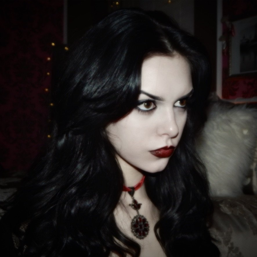 A woman with long black hair and dark eye makeup, wearing red lipstick, a red necklace with a pendant, and a black top, sitting on a couch with a pillow, in a dimly lit room.