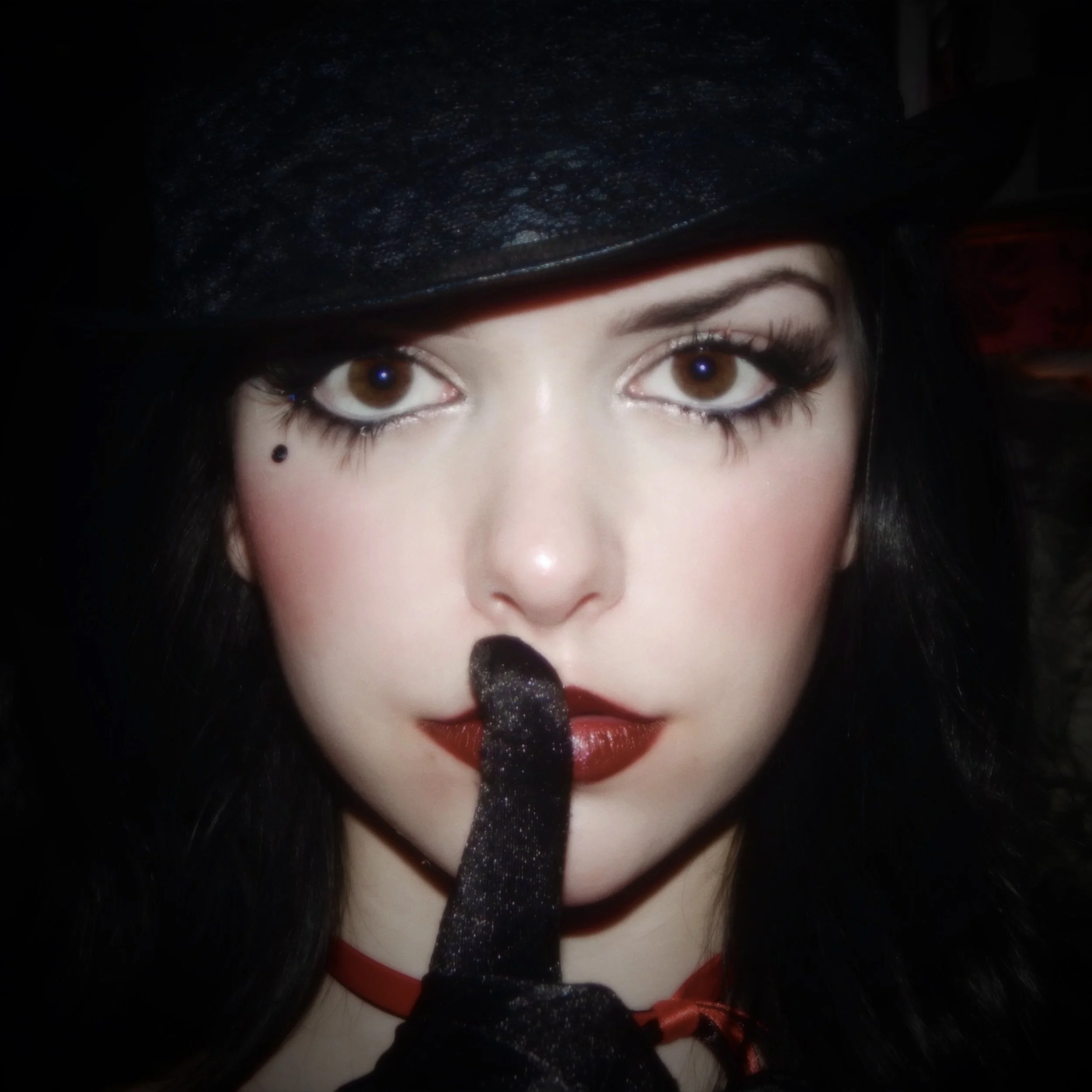 Close-up of a woman with dark hair, wearing a black lace hat and a black glove, holding her finger with red lipstick to her lips in a shushing gesture.