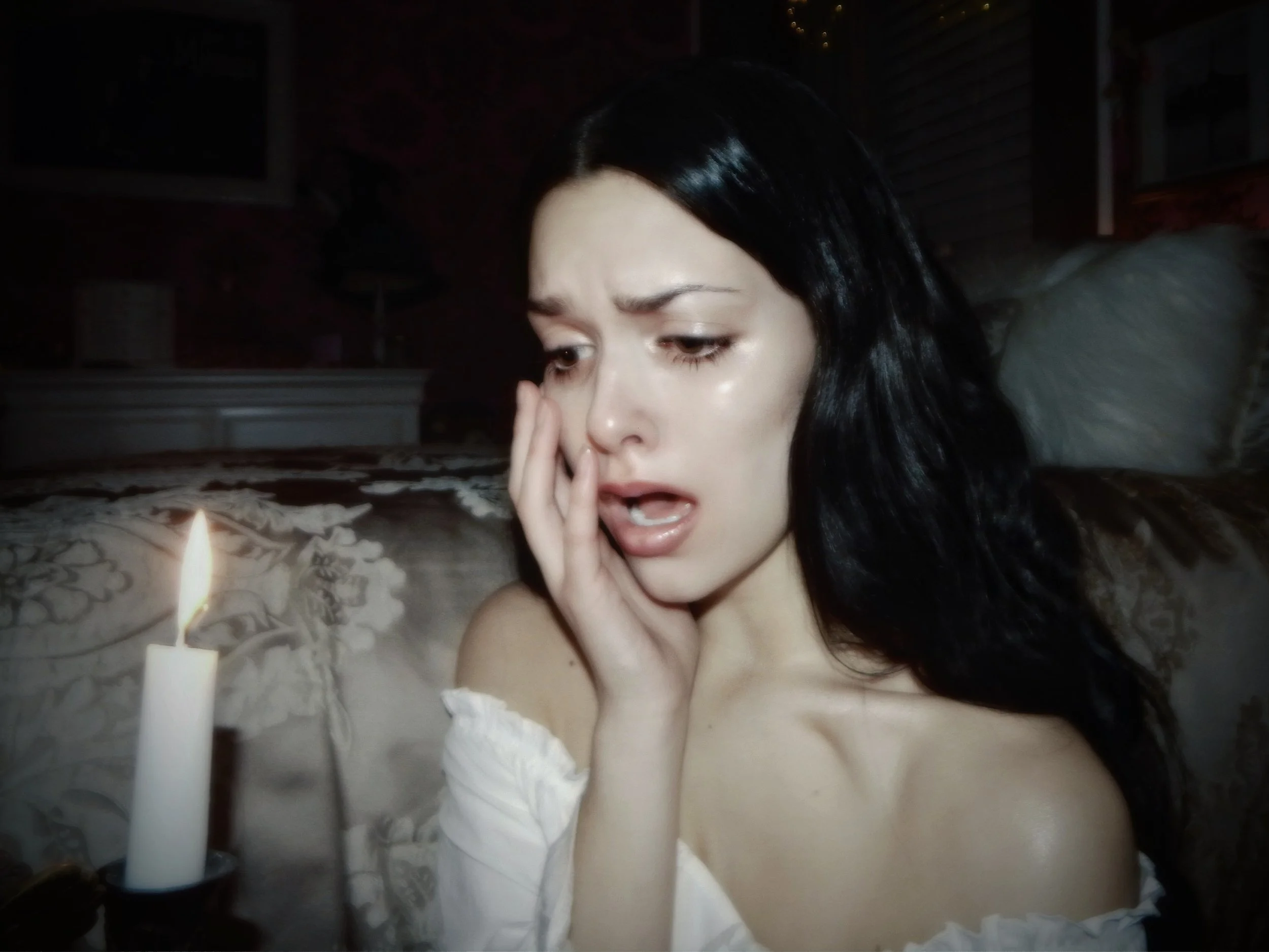 A woman with long black hair and fair skin appears distressed, holding her face, with a lit candle in front of her.
