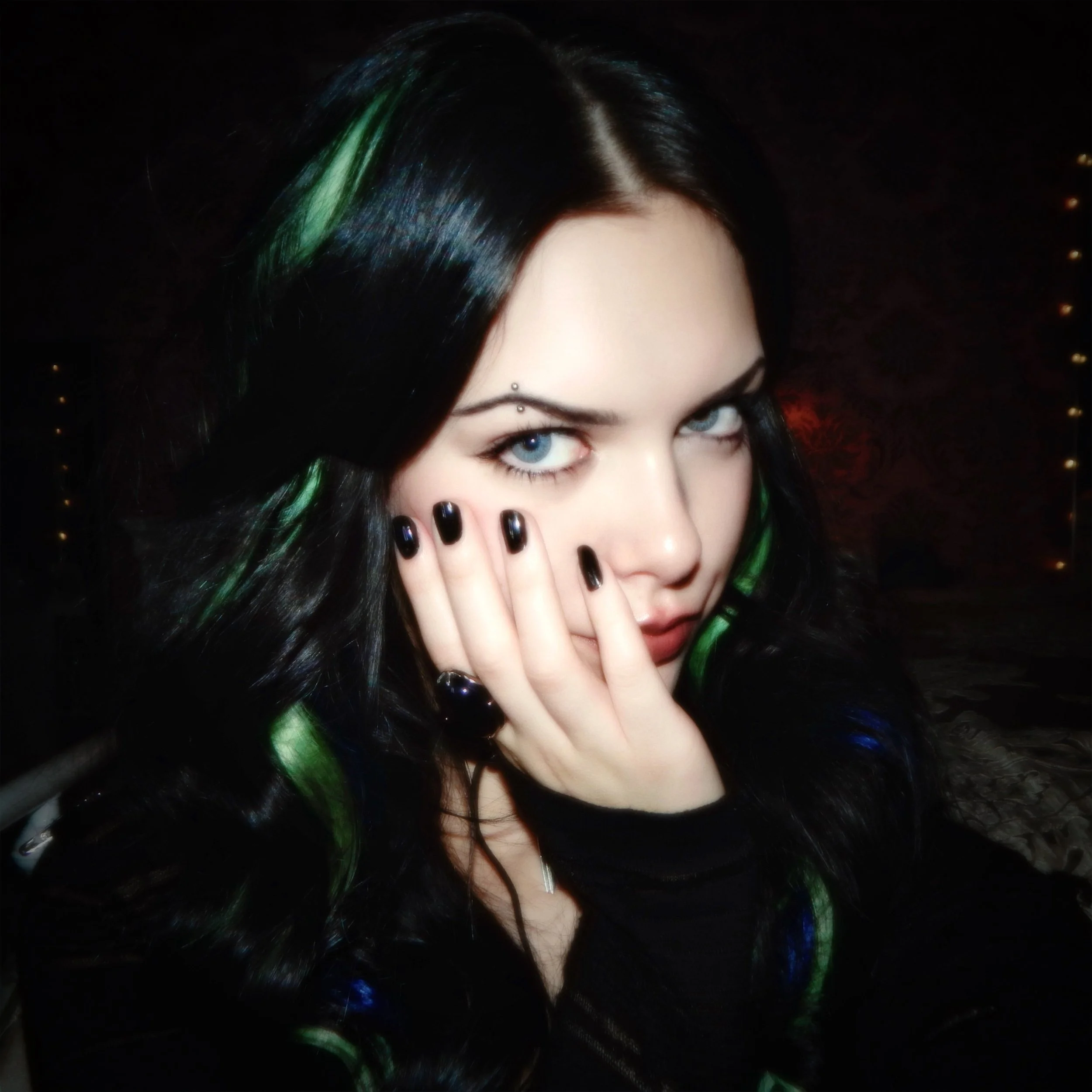 A woman with striking blue eyes and long black hair with green highlights covers part of her face with her hand, revealing black painted nails, intense makeup, and a small decorative piercing above her eyebrow, in a dark setting.