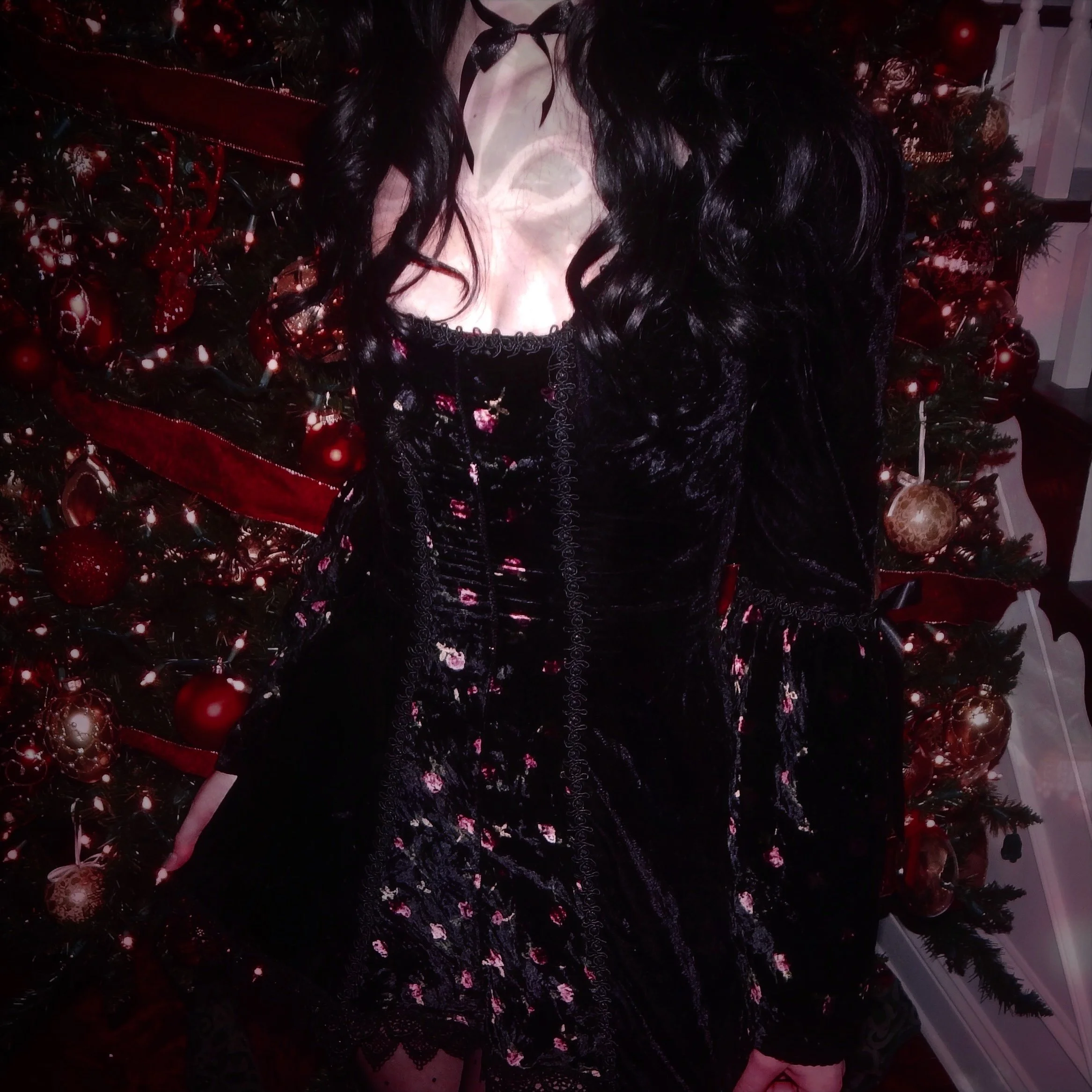 Person in a black dress with pink floral patterns standing in front of a decorated Christmas tree with red and gold ornaments.