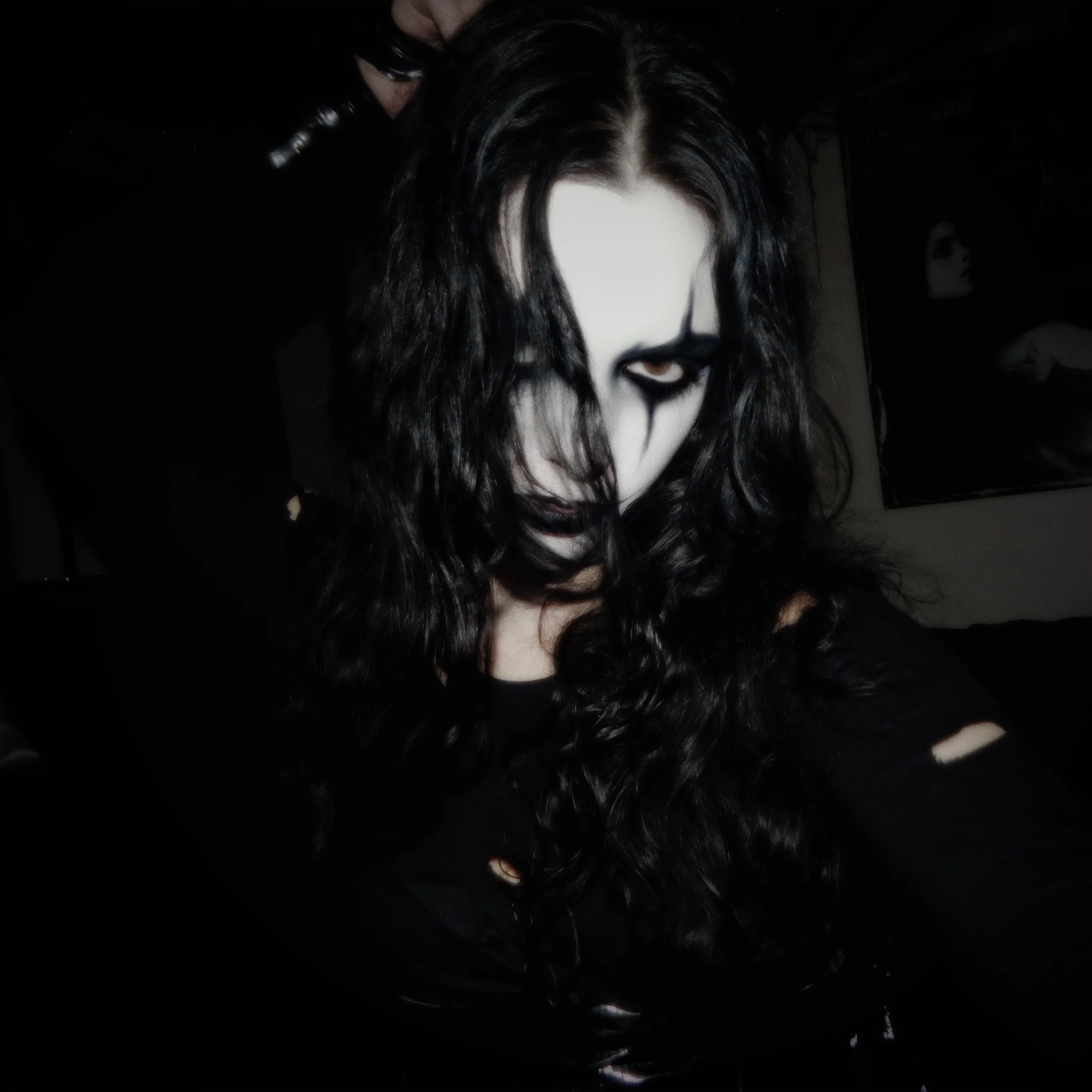 Person with black curly hair, white face makeup, dark eye makeup, and black face paint design, wearing a black shirt with ripped sleeves, in a dark room.