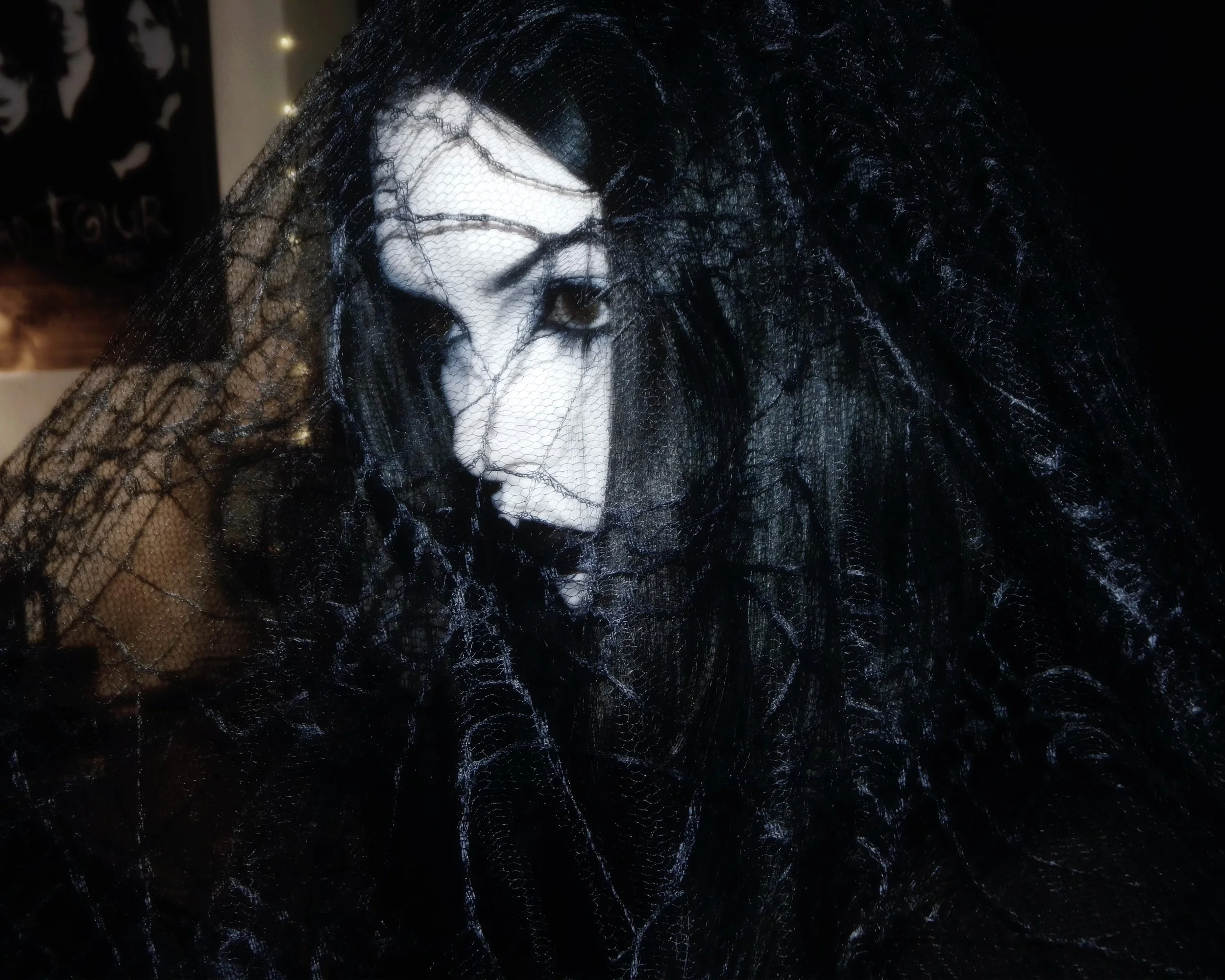 A person with pale makeup, dark hair, and black lipstick, wrapped in black spiderweb-like fabric, with a gothic aesthetic.