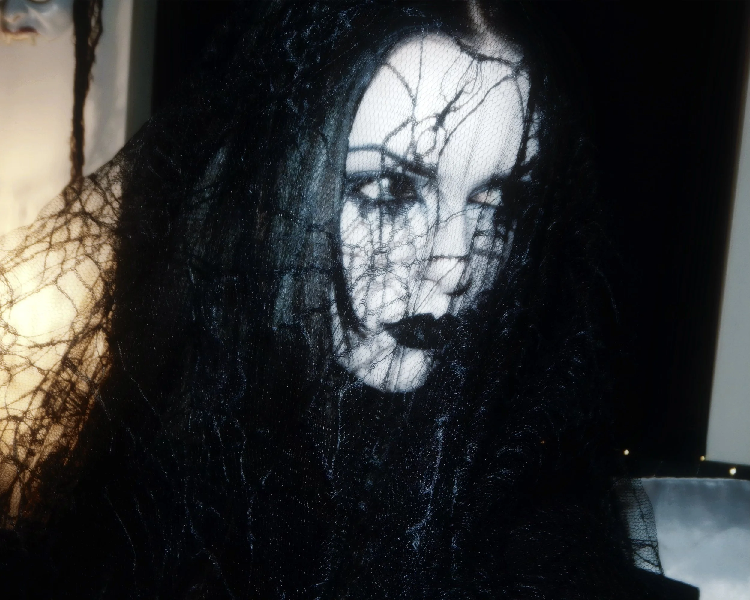 A woman with pale makeup and dark eye makeup, wearing a black veil and lace, appears to be in a dark setting, giving a mysterious and gothic appearance.