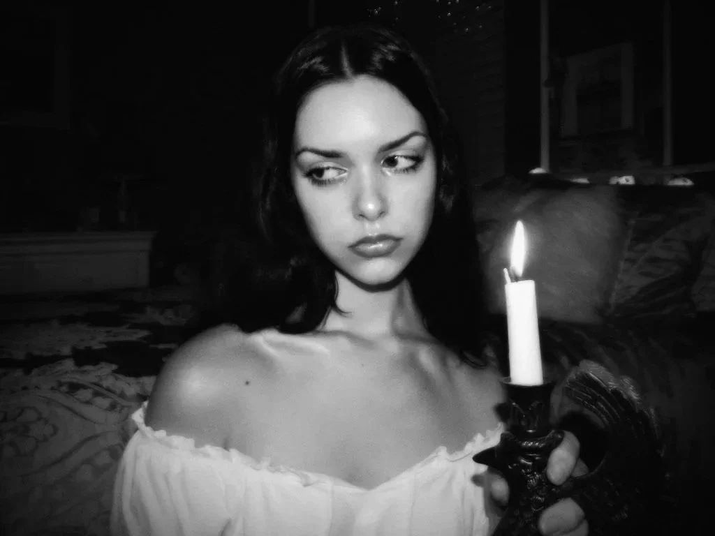 Black and white photo of a woman with dark hair holding a lit candle in a decorative holder, looking to the side with a serious expression, in a dimly lit room.