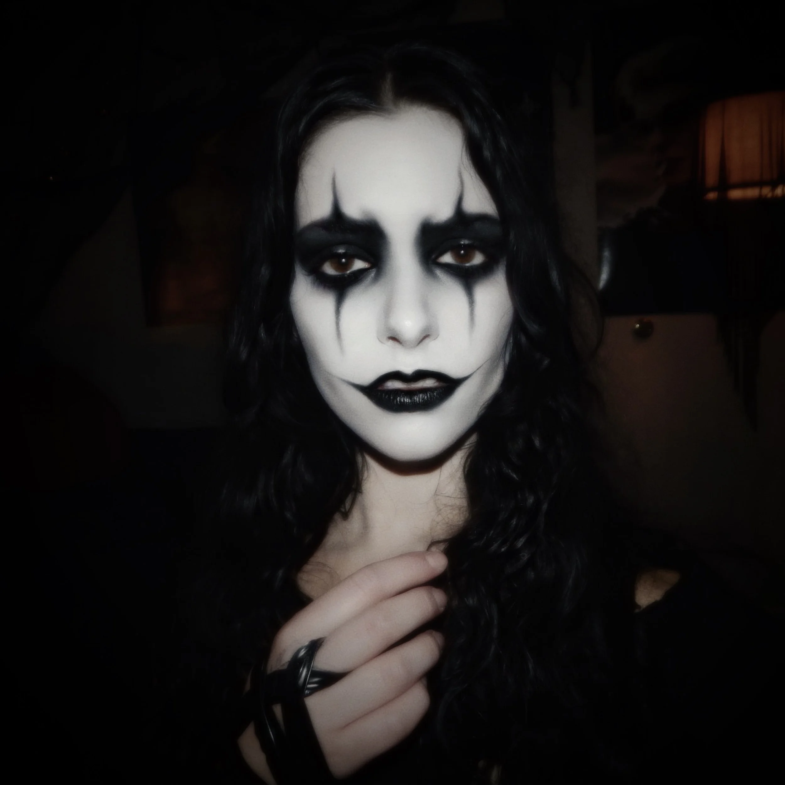 A woman with black hair and gothic makeup, including dark eye makeup, black lipstick, and white face paint, looking directly at the camera.