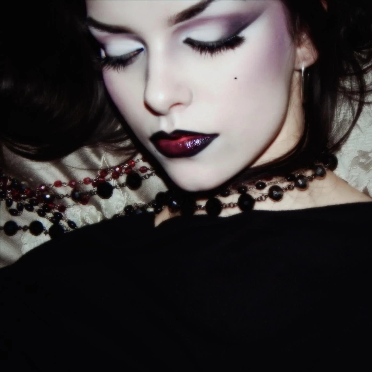 Close-up of a woman with dark hair, goth makeup, dark lipstick, and long eyelashes, lying with her eyes closed, wearing a black necklace and earrings.
