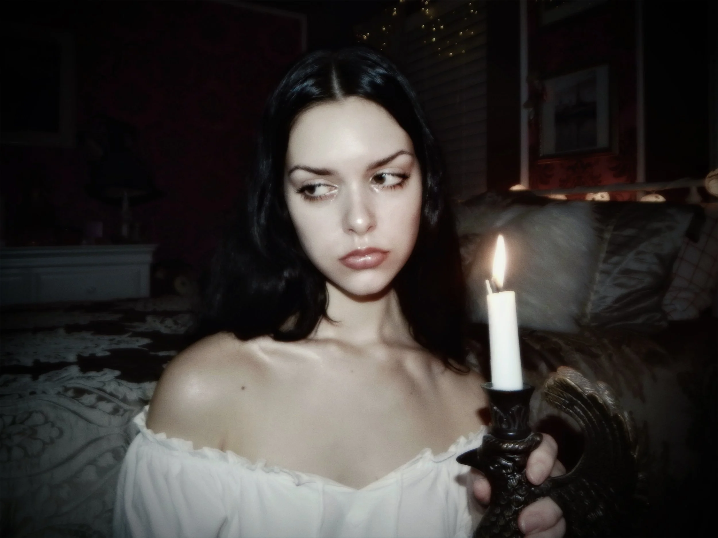A woman with dark hair holding an unlit candle in a candle holder in a dimly lit room.