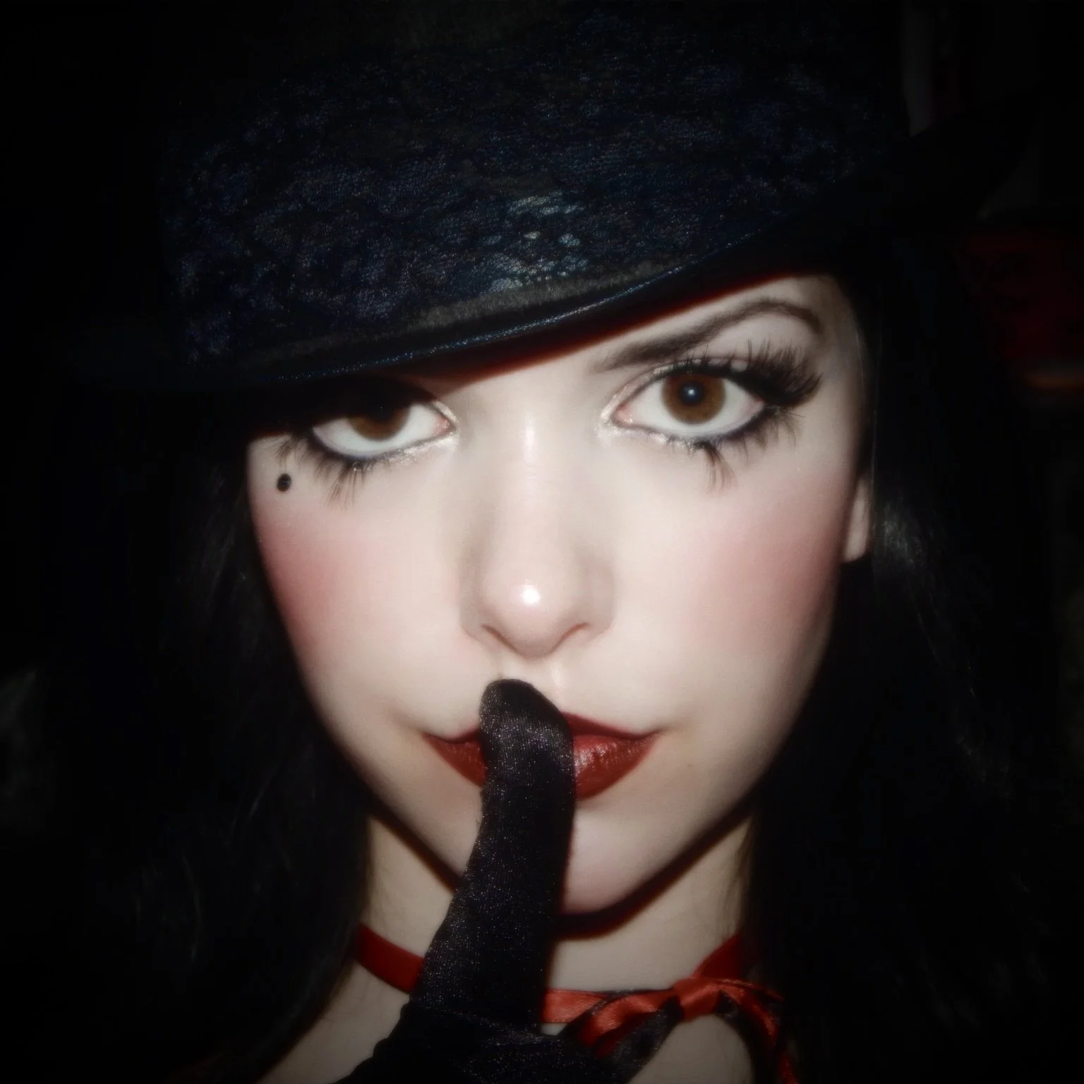 A woman with brown eyes, dark hair, wearing a black hat and gloves, and red lipstick, is holding her index finger to her lips in a shushing gesture.