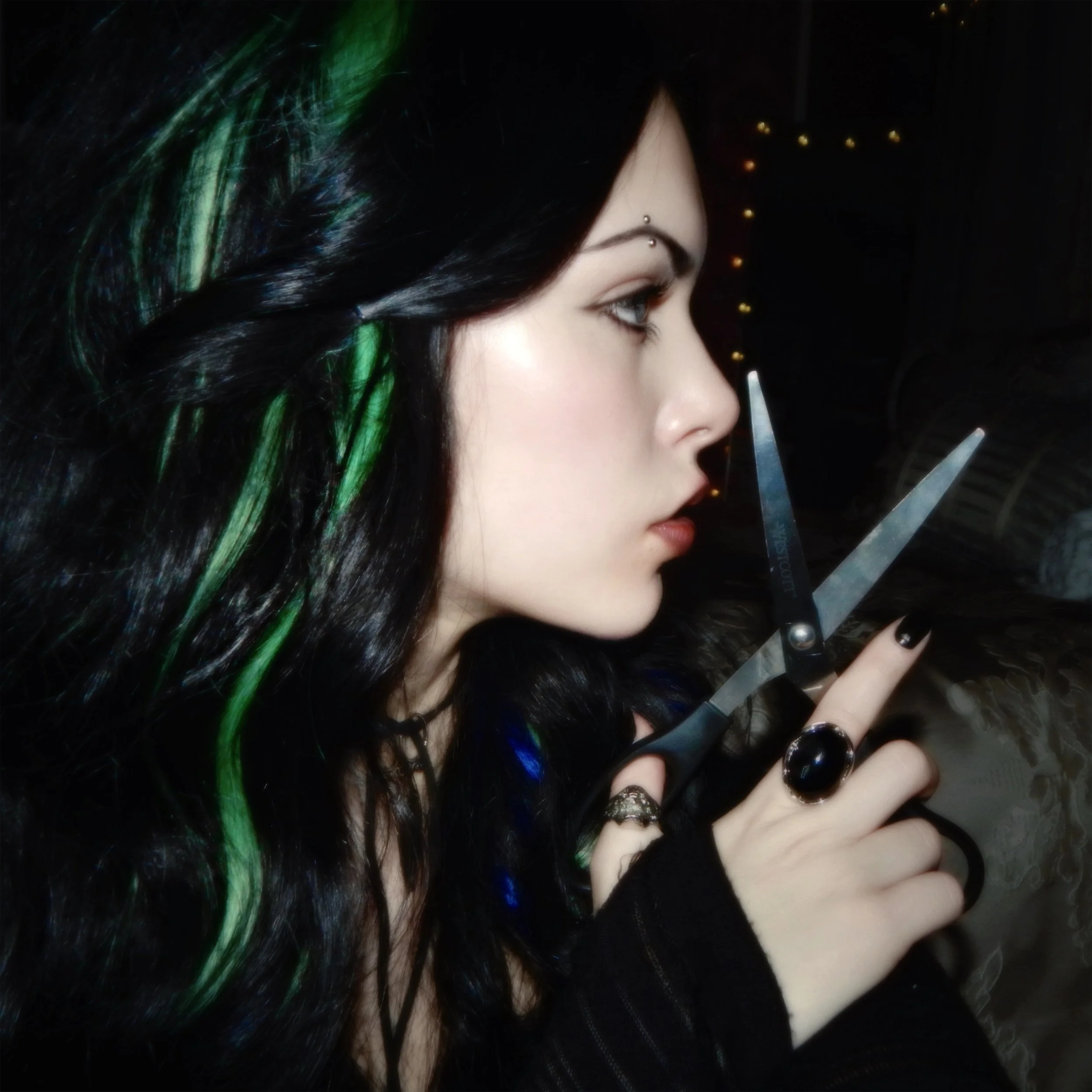 A woman with black hair and green streaks, holding scissors near her face, with dark clothing and jewelry, in a dimly lit room with fairy lights.