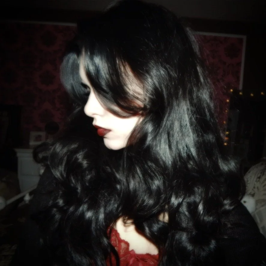 A woman with long, wavy black hair and red lipstick, looking down, dressed in black and red, in a dimly lit room with patterned wallpaper.