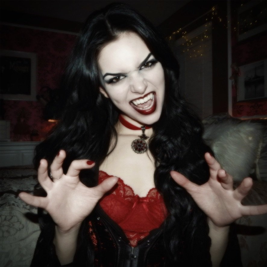 A woman with long black hair making a fierce expression with her mouth open and hands raised, dressed in red and black gothic style with dark makeup in a room with dim lighting.