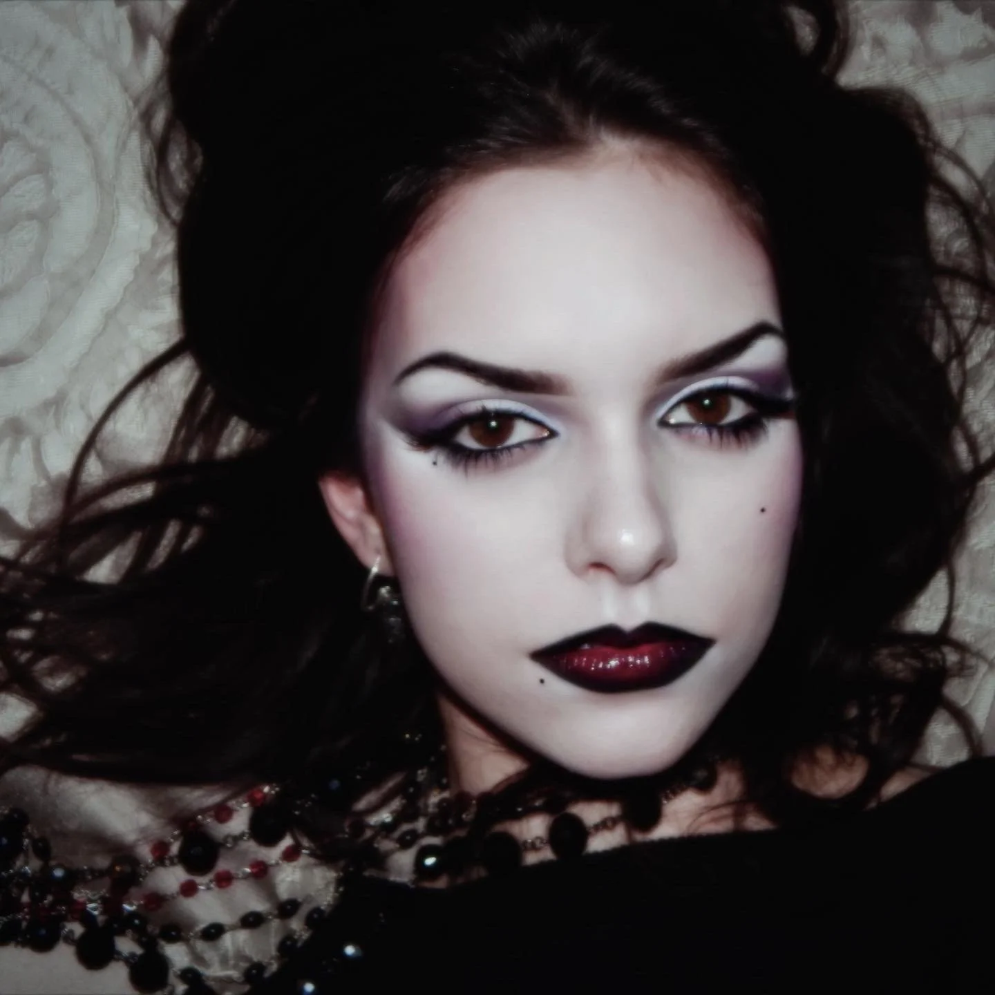 A woman with dark hair and dramatic makeup lying down on a patterned surface, wearing dark lipstick and jewelry.