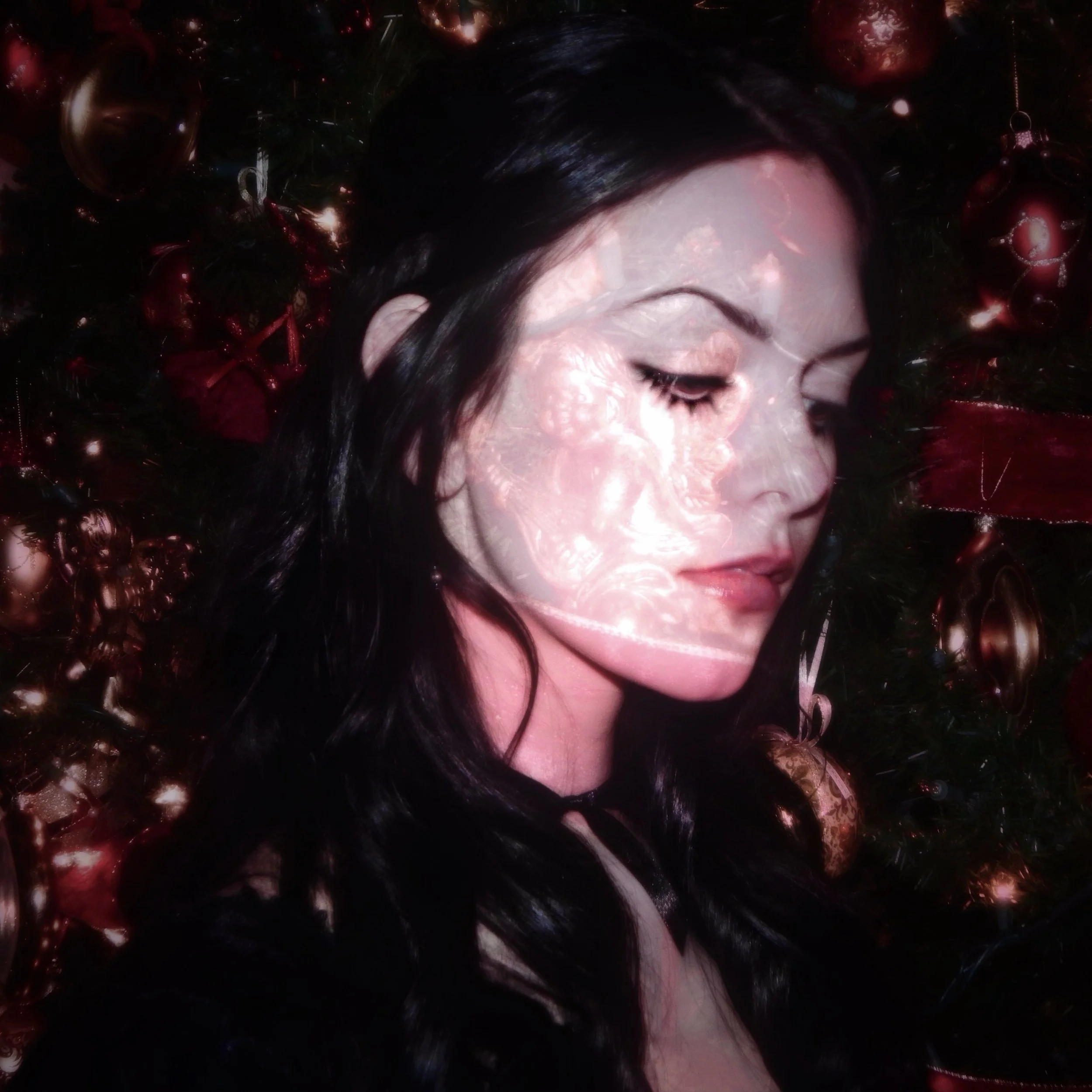 A woman with dark wavy hair, closed eyes, and light makeup, surrounded by red and gold Christmas ornaments and decorations.