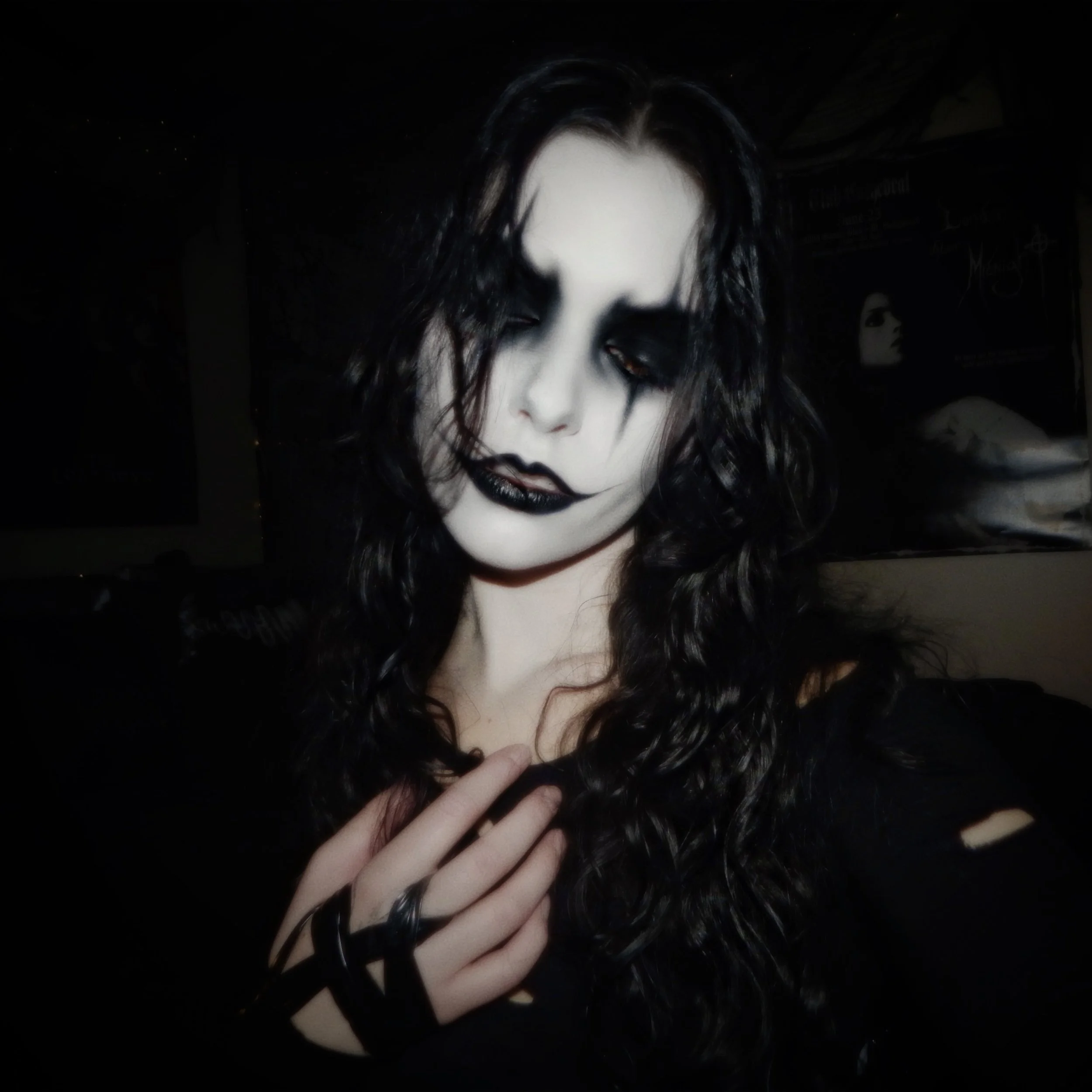 Person with black curly hair, white face makeup, black lipstick, and dark eye makeup, posing with a hand on chest in a dark room.