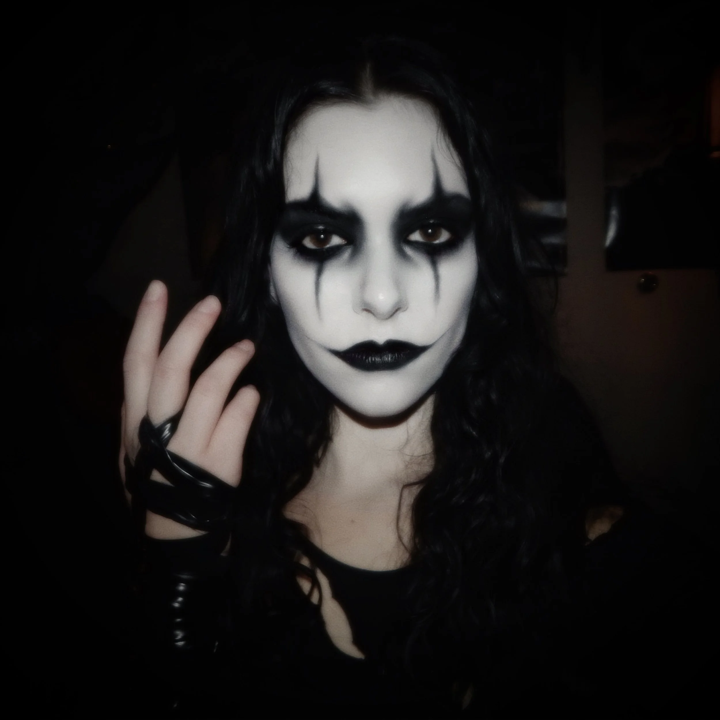 Person with black hair and heavy black makeup, including dark lipstick and black face paint resembling a clown or gothic style, holding their hand near their face with fingers adorned with black accessories, standing in a dimly lit room.