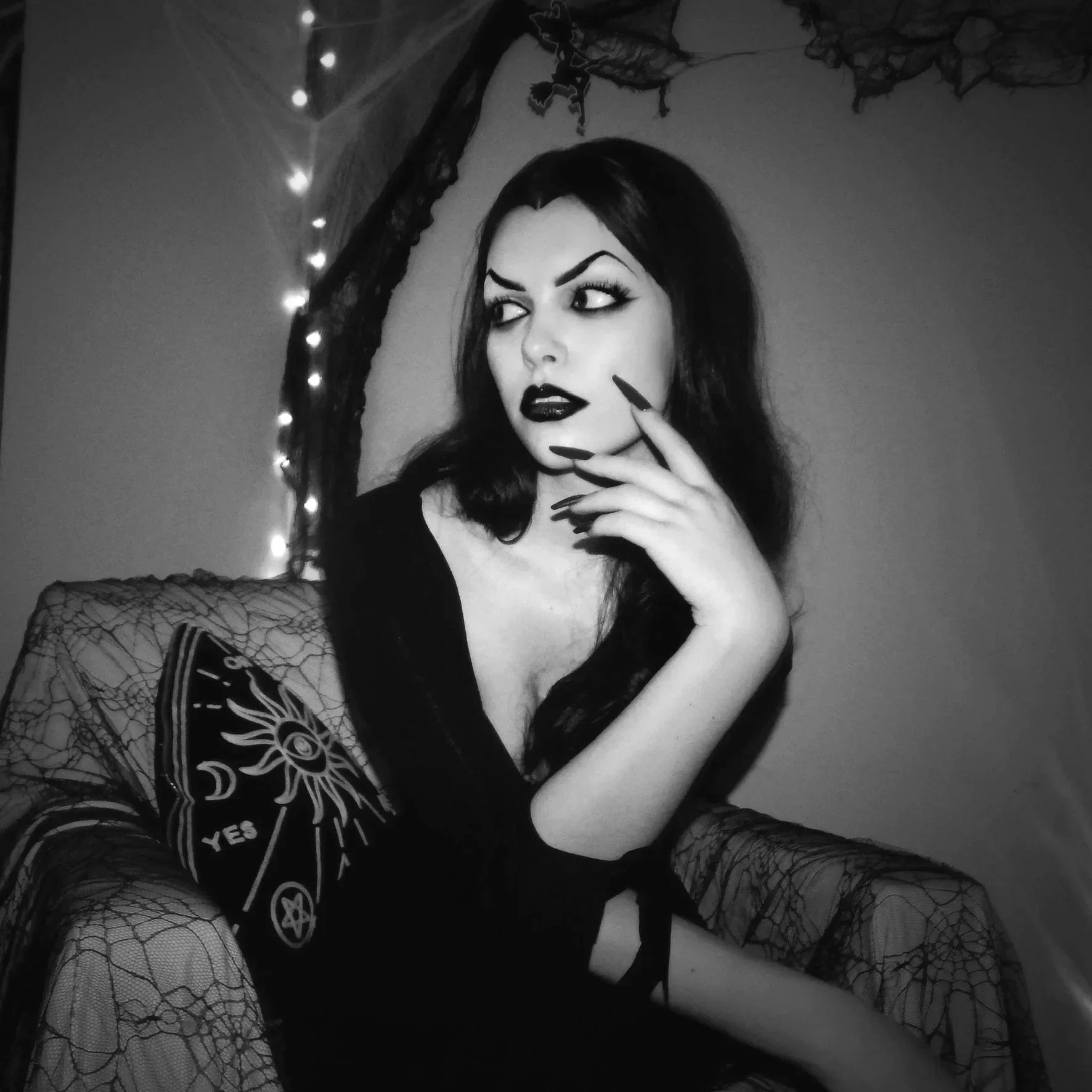 A black and white photo of a woman with dark hair and makeup, sitting on a gothic-style chair, with her hand near her face, surrounded by dark drapery and string lights.