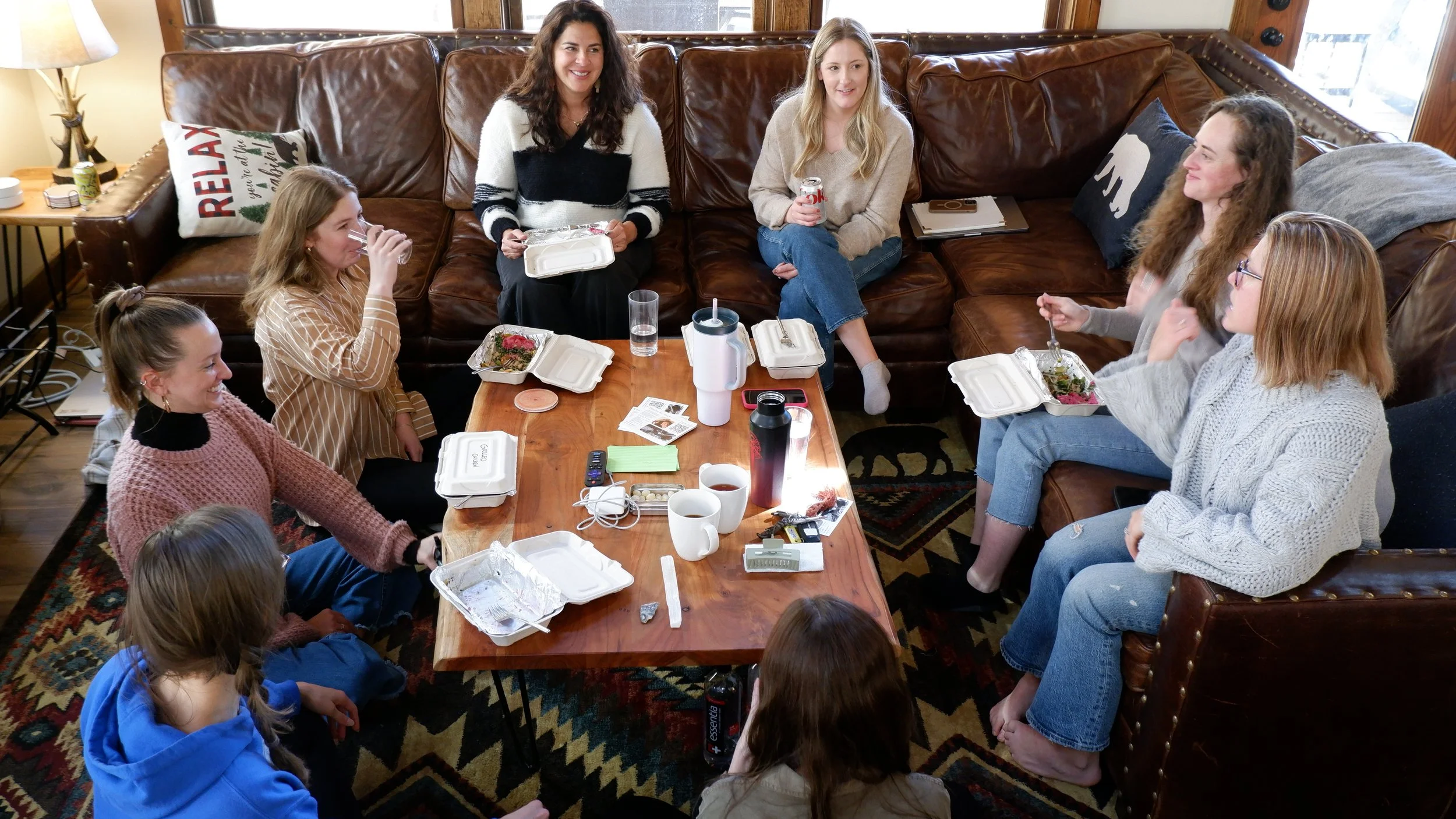 Group of women and children gathered in a cozy living room, sitting around a wooden table with food containers, drinks, and personal items, engaging in conversation.