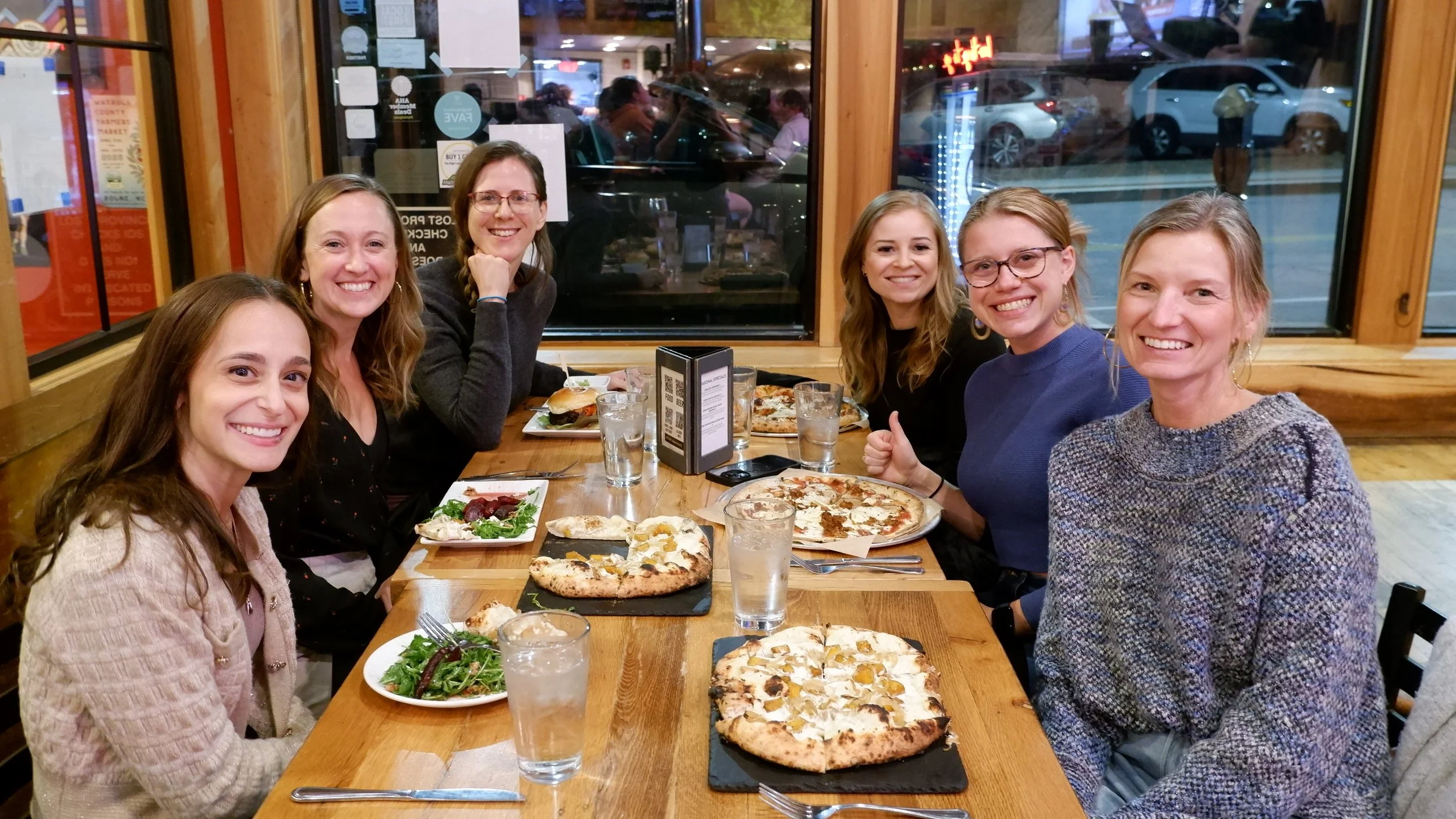 Five women sitting around a restaurant table enjoying pizza and salads, smiling at the camera inside a cozy restaurant with wooden decor and large windows.