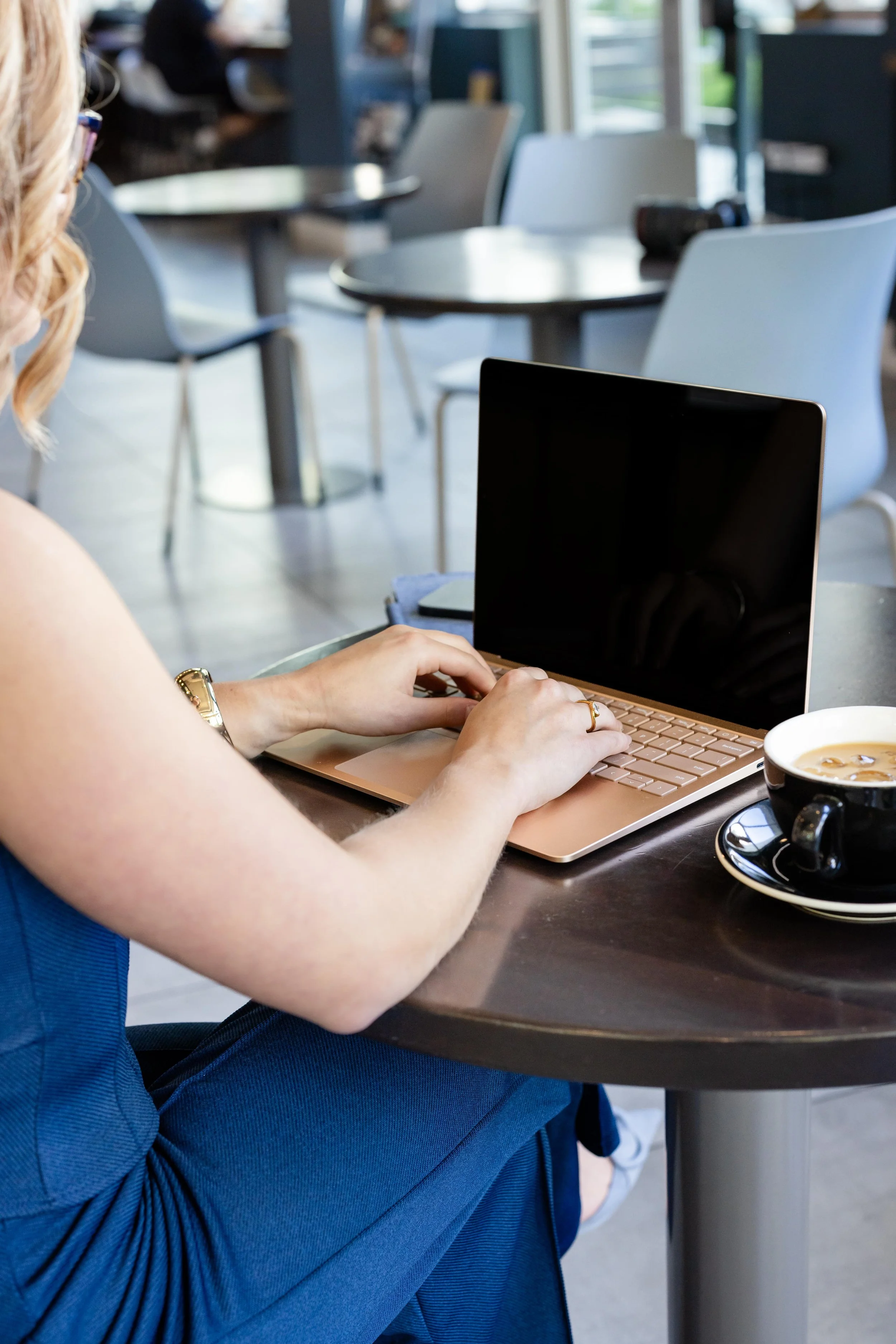 A woman typing on a pink laptop at a cafe table with a cup of coffee nearby.