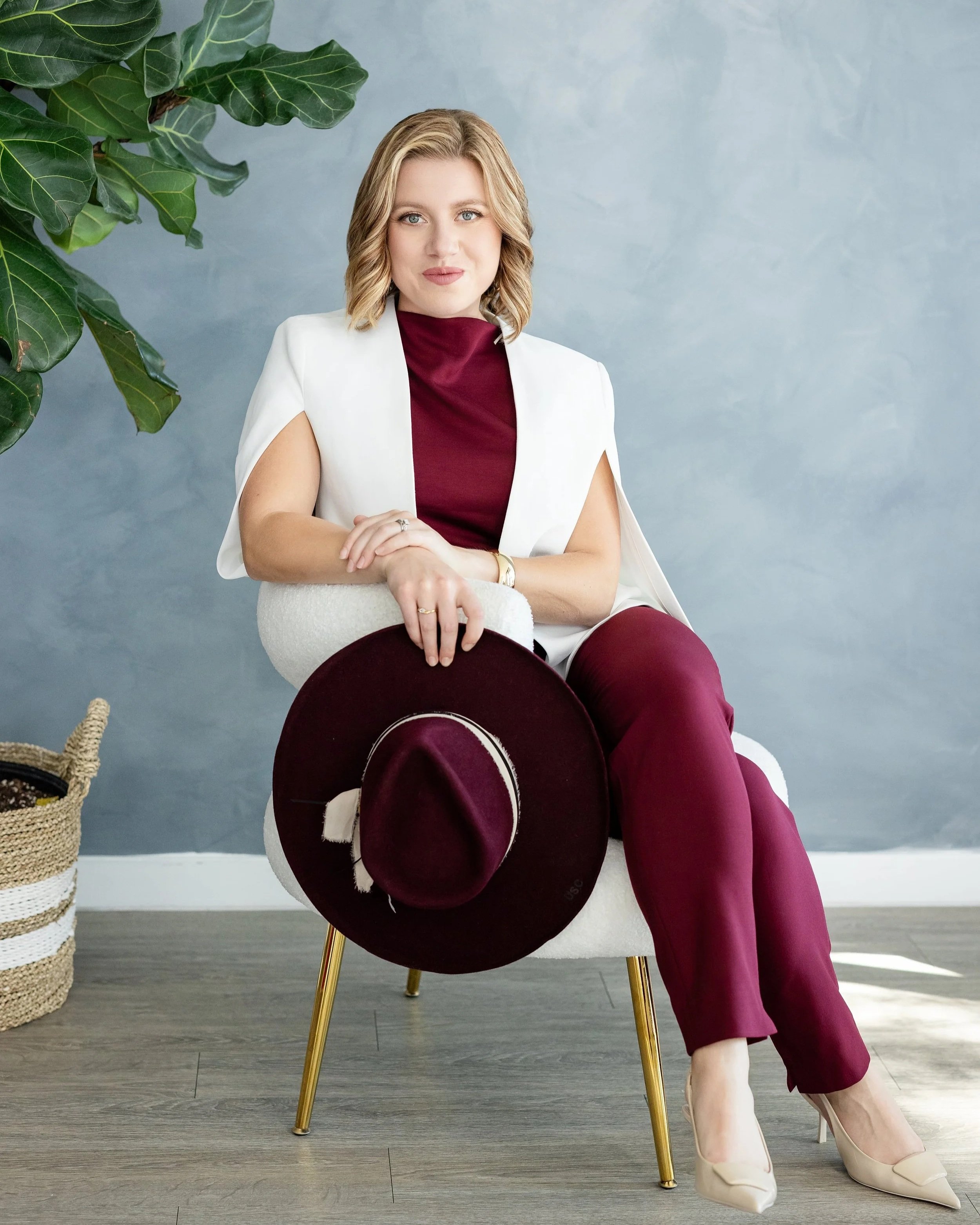 A woman with shoulder-length blonde hair sitting on a white chair with gold legs, holding a burgundy hat. She is wearing a burgundy top, matching pants, a white sleeveless blazer, and beige high heels. There is a large green plant on the left and a woven basket on the floor. The background is a blue-gray wall.