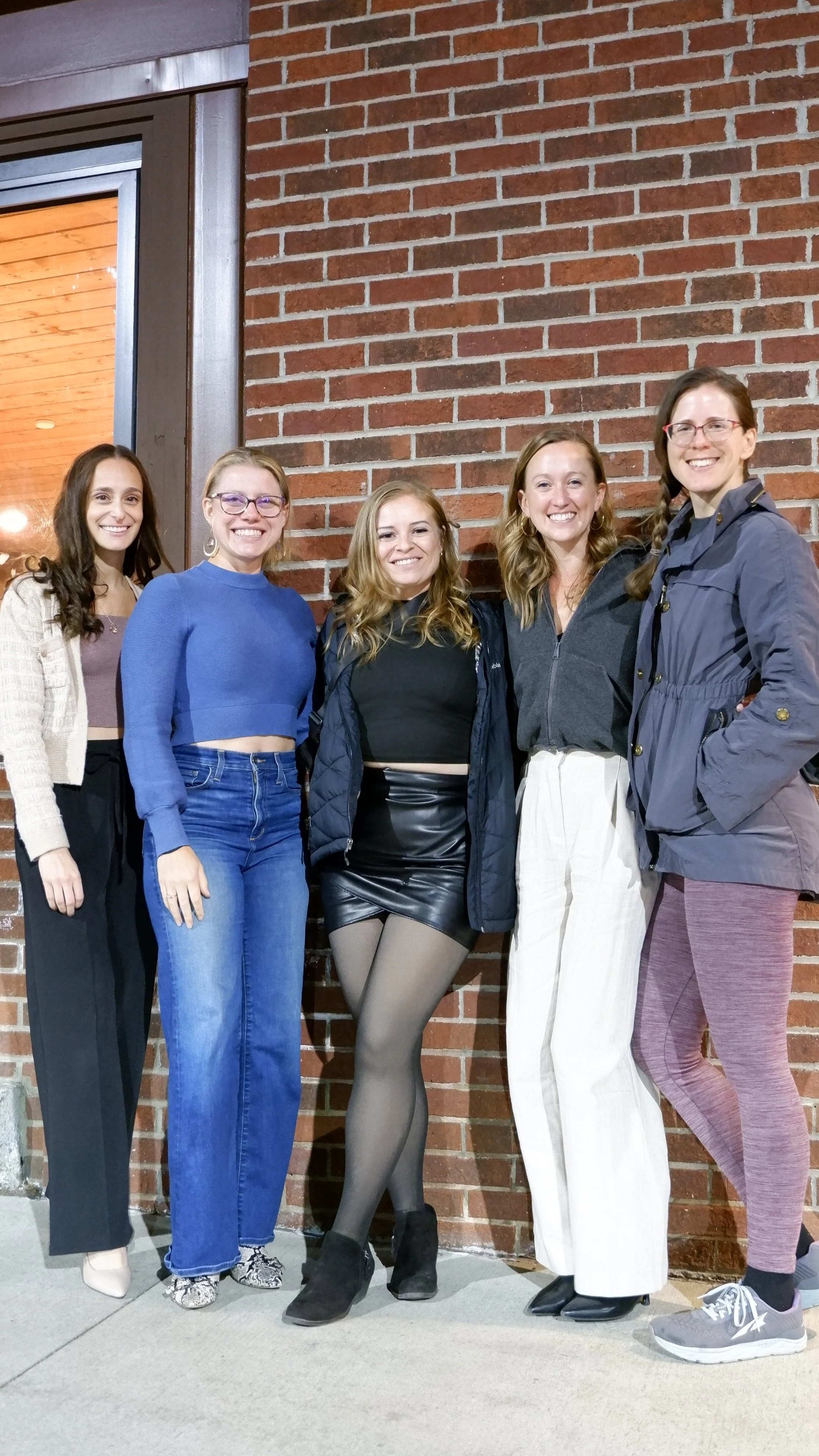 Five women smiling and standing in front of a brick wall. They are dressed casually and appear to be indoors, near a window or glass door.