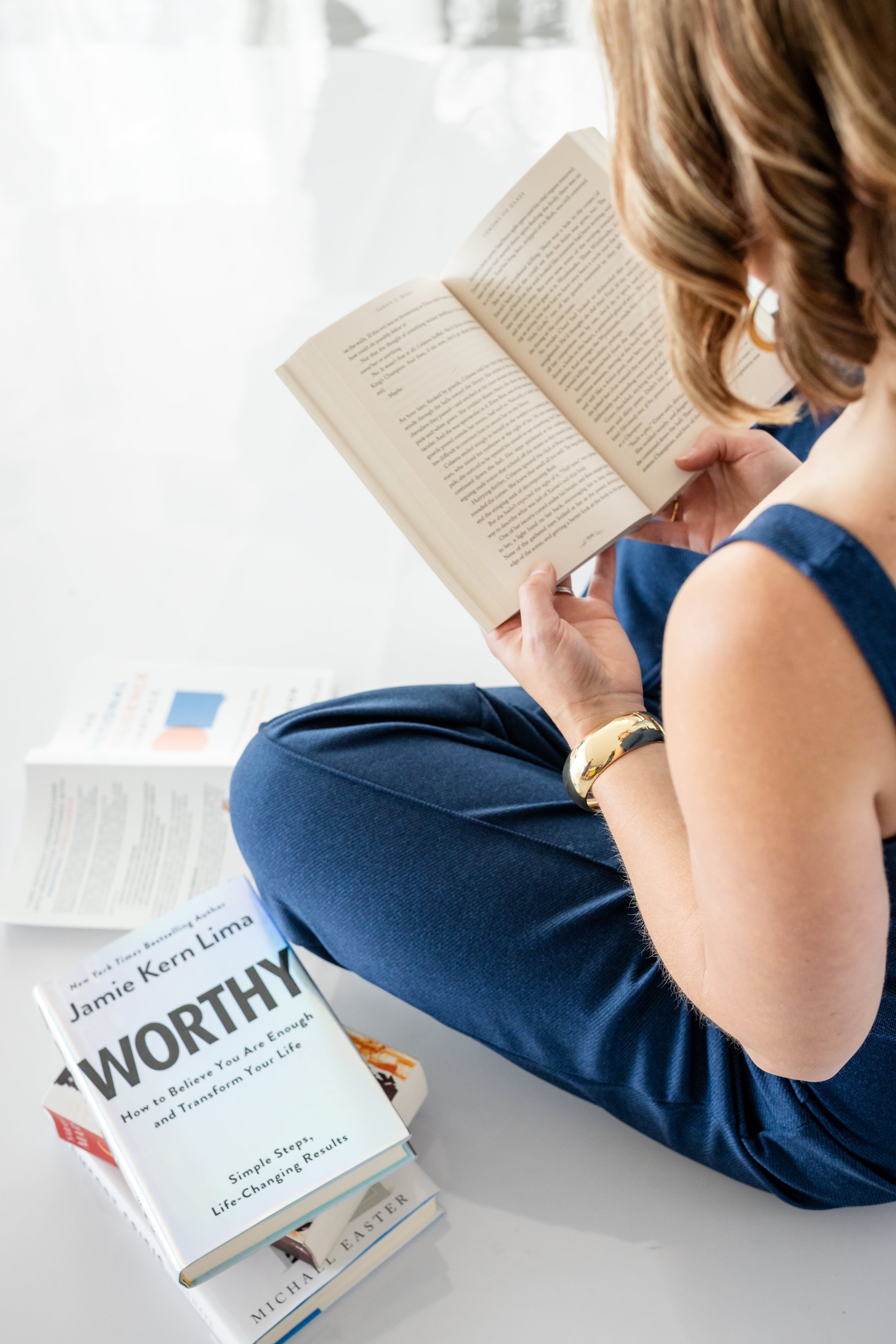 A person sitting on the floor, reading a book. On the floor next to them are several books, including one titled 'WORTHY' by Jamie Kern Lima.