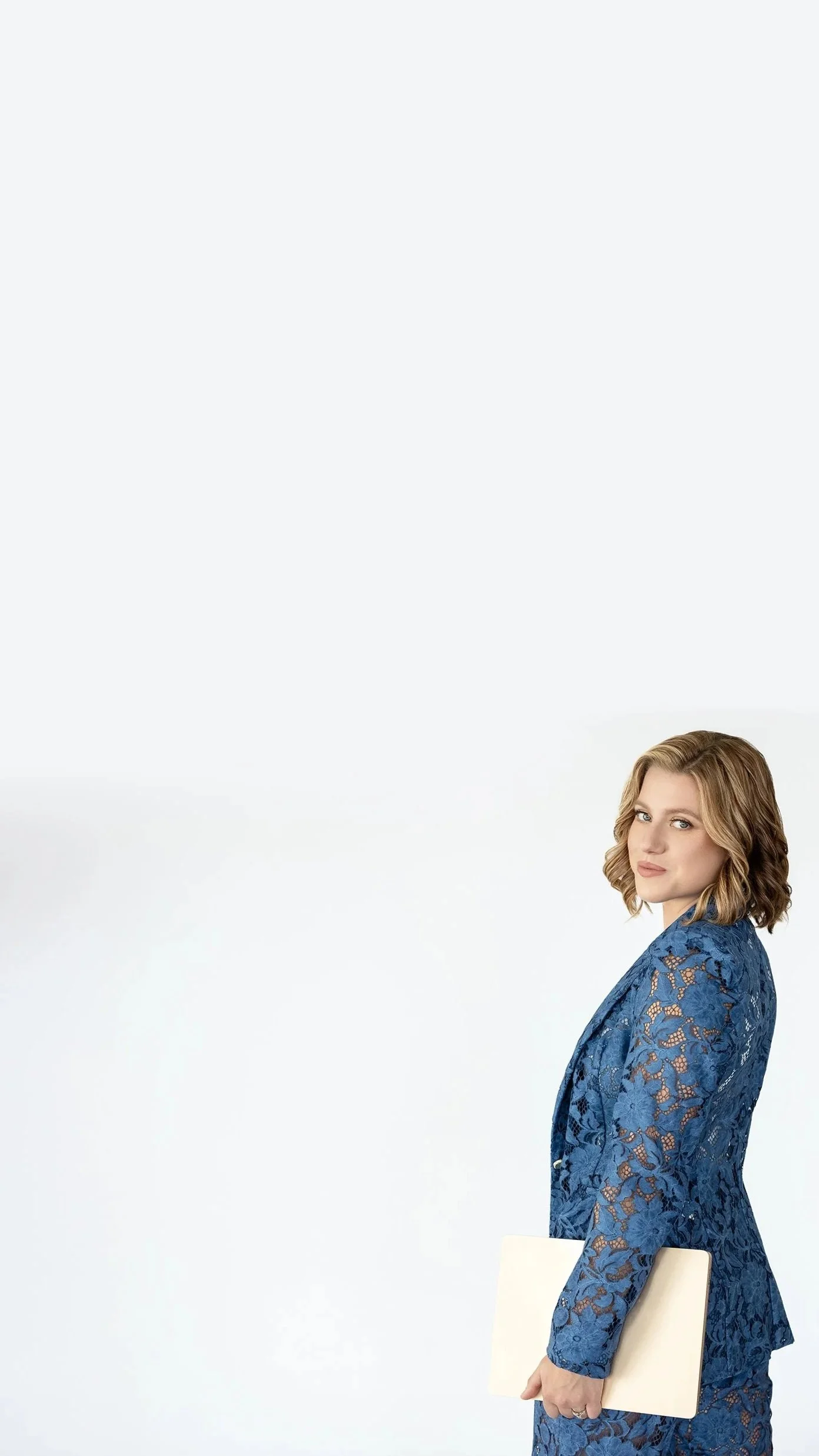 A woman with shoulder-length blonde hair and light makeup holding a closed beige laptop, wearing a blue lace blazer and matching skirt, standing against a plain white background.