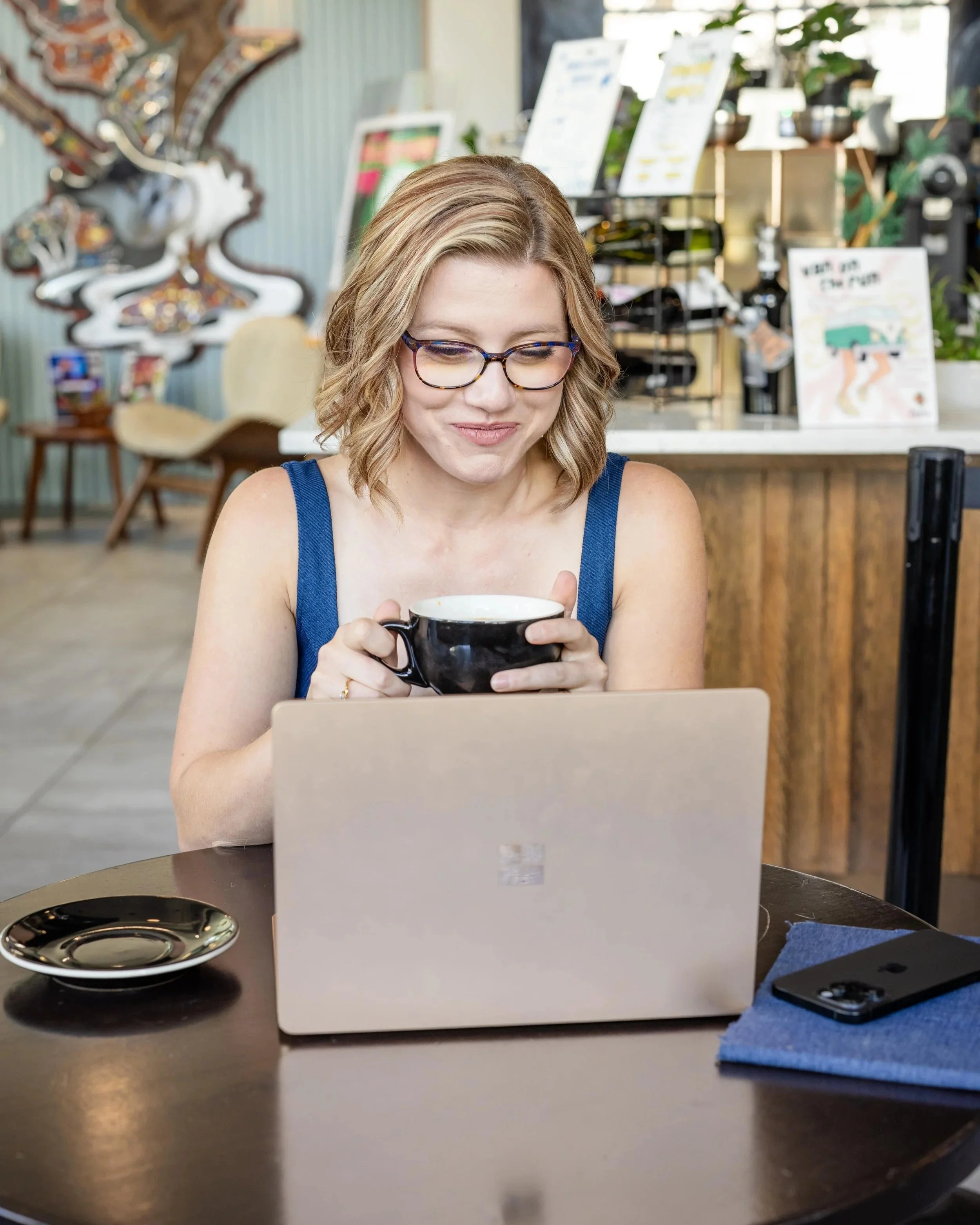 A woman with blonde wavy hair and glasses sits at a table in a cafe, holding a black mug and looking at her laptop screen.