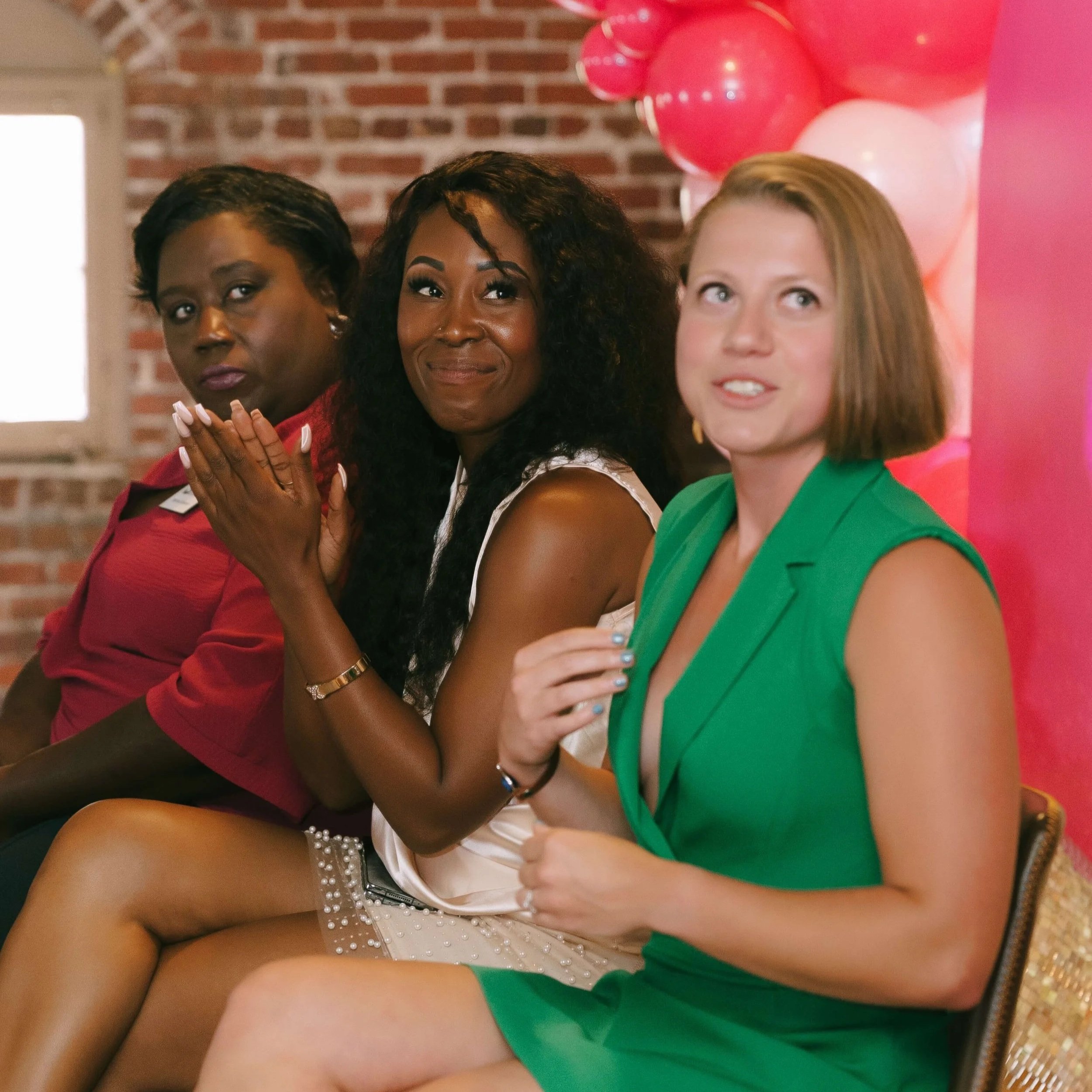 Three women sitting side by side at a celebration event with pink and red balloons in the background.