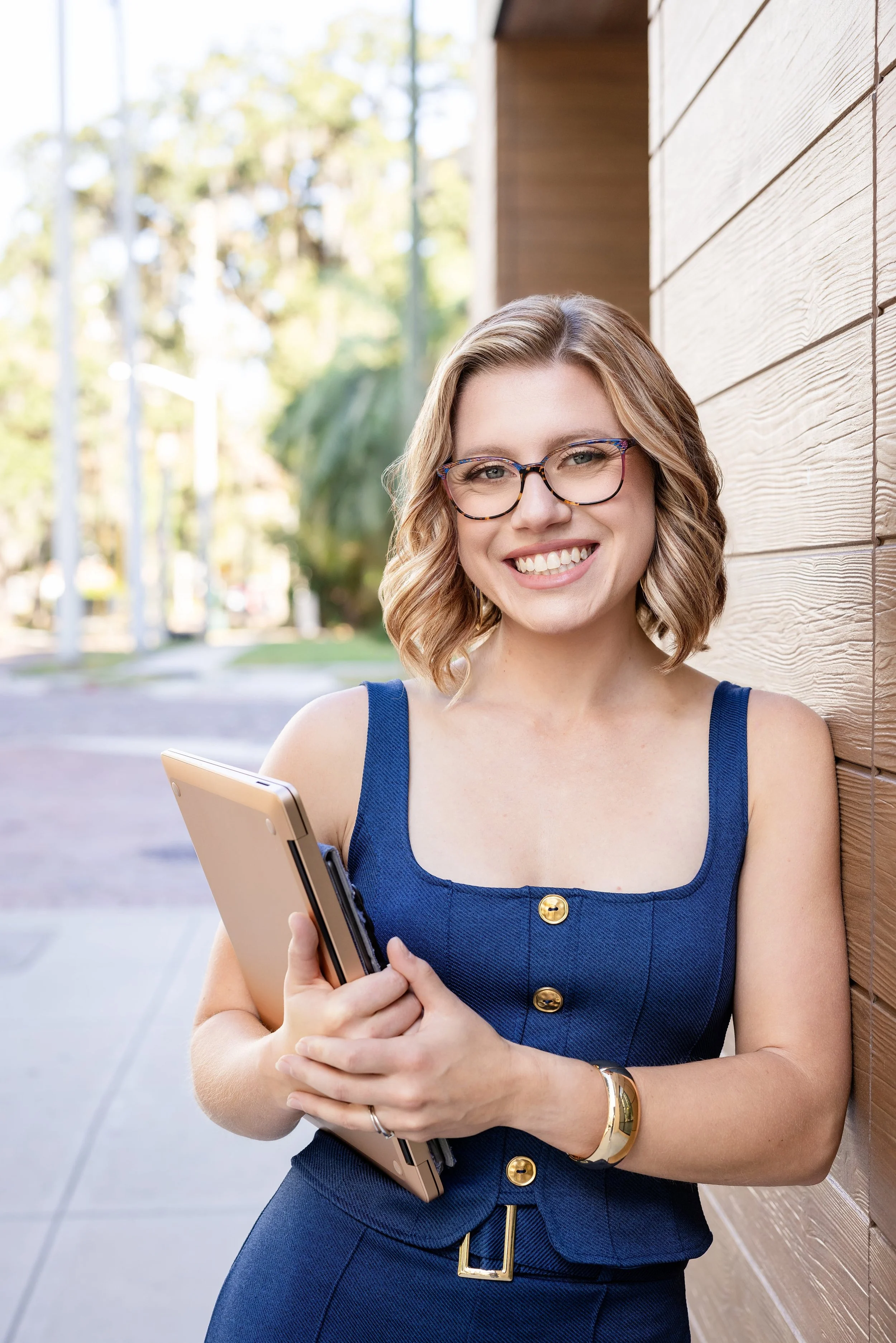 A smiling woman with glasses standing outdoors, holding a tablet, and leaning against a wood-paneled wall.