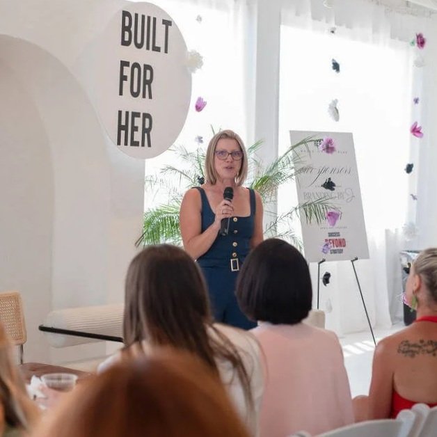 A woman speaking into a microphone at an indoor event with a sign that reads 'Built For Her' in the background and an audience of women seated and listening.