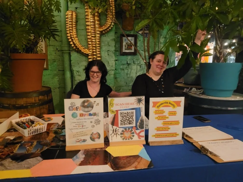 Two women smiling and sitting behind a table at an indoor event or market, with signs, a QR code, and items displayed on the table. One woman is reaching up toward a potted plant.