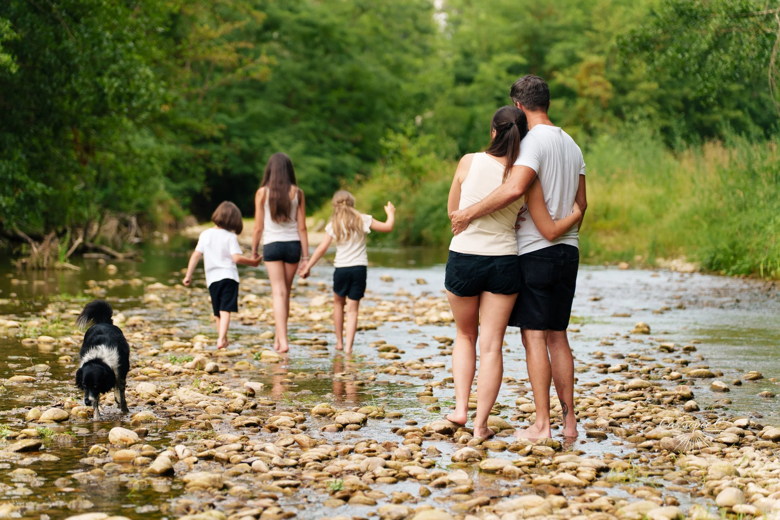 Un groupe de personnes, dont des enfants, se promènent pieds nus dans une rivière au milieu d'une forêt verdoyante, avec un chien noir et blanc qui explore l'eau.