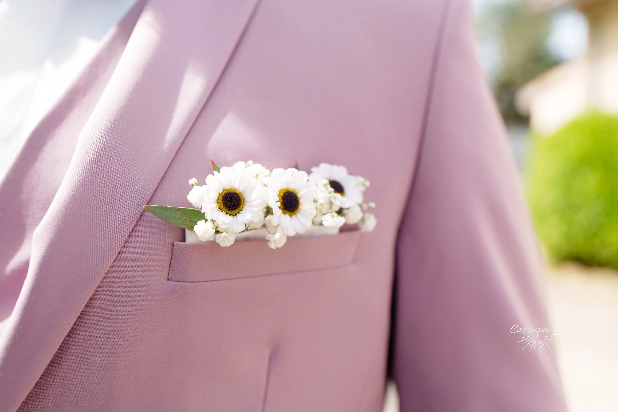 Une personne portant une veste de costume rose clair avec une poche de poitrine contenant un bouquet de petites fleurs blanches avec un centre foncé et quelques feuilles vertes.